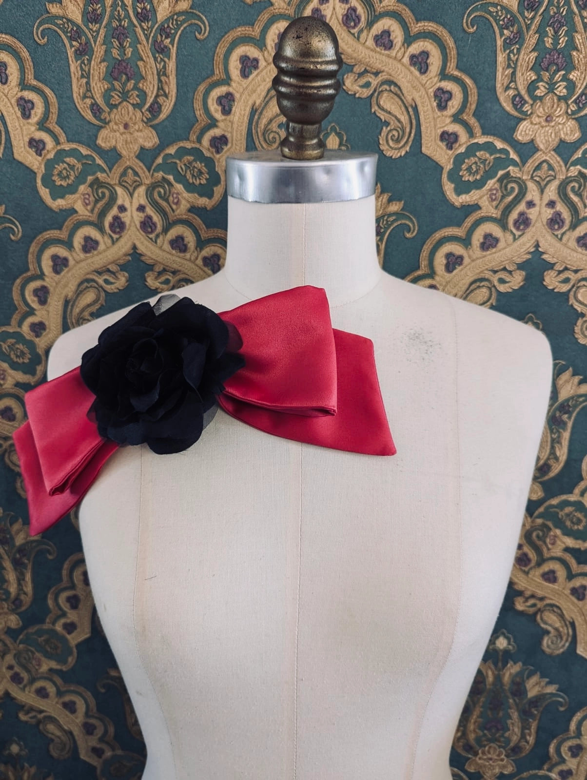 Broochella viva l'amor handmade pink satin bow brooch with black flower centre.  It is pinned to a mannequin. The background is green and gold damask wallpaper.