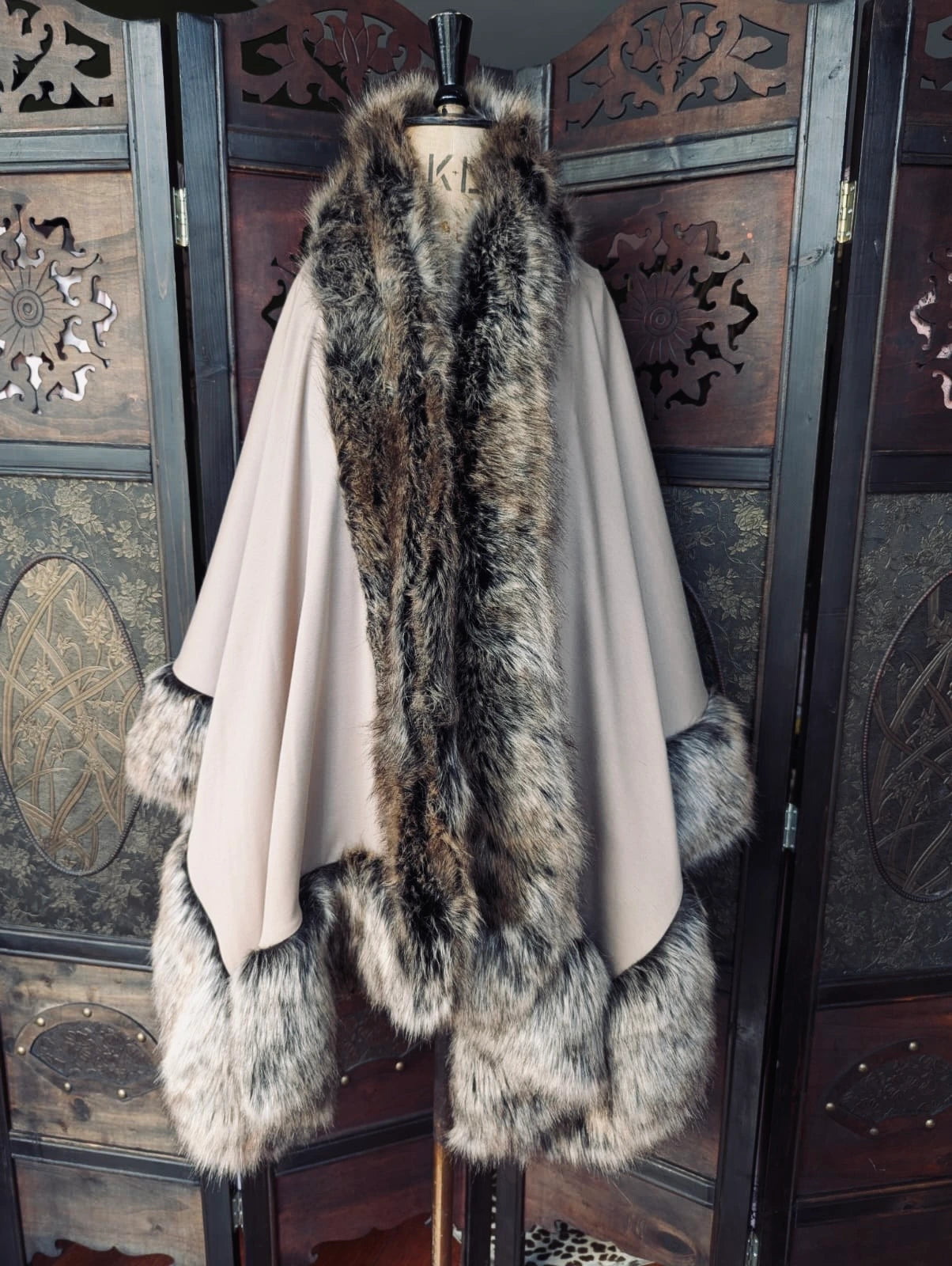 A Broochella Stylist's Closet Donna Salyers Fabulous Furs OSFA beige cape with a large dark brown and gray faux  fur collar, cuffs, and hem, displayed on a mannequin in front of a dark wooden decorative screen.