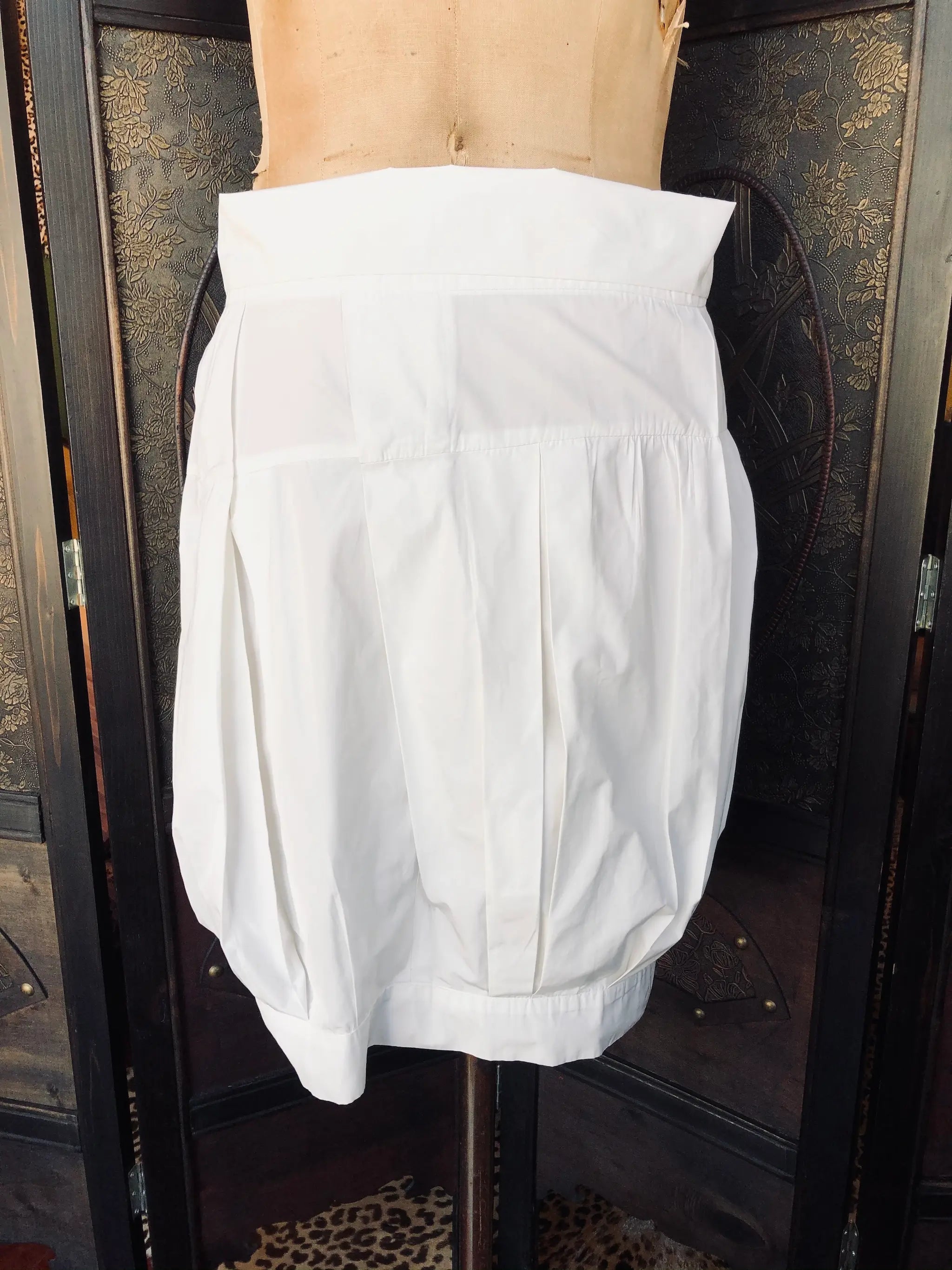 Broochella Dries Van Noten 36 100% Polyester, White, Sculptural cocoon silhouette, with soft pleating and panel detailing. it is pictured on a vintage mannequin. The background is an ornate, vintage, dark wood room divider. 