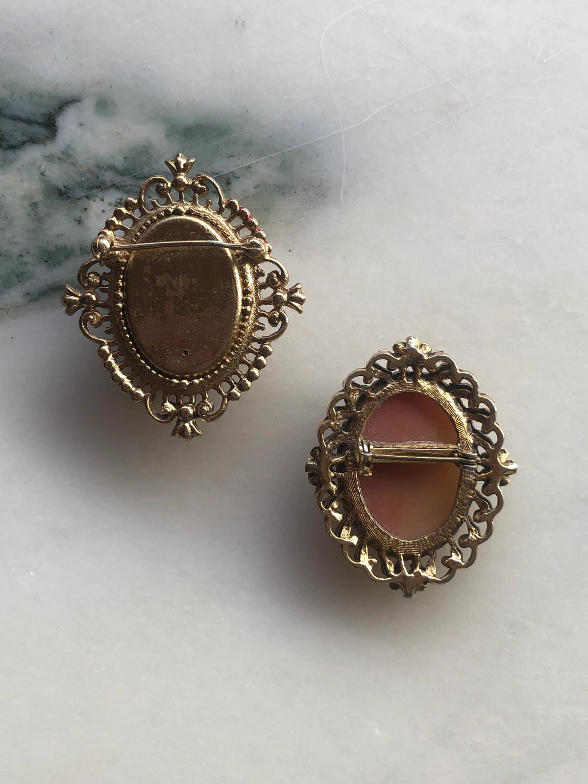 The back view of Two Broochella Duo Sorelle vintage cameo brooches on a marble surface