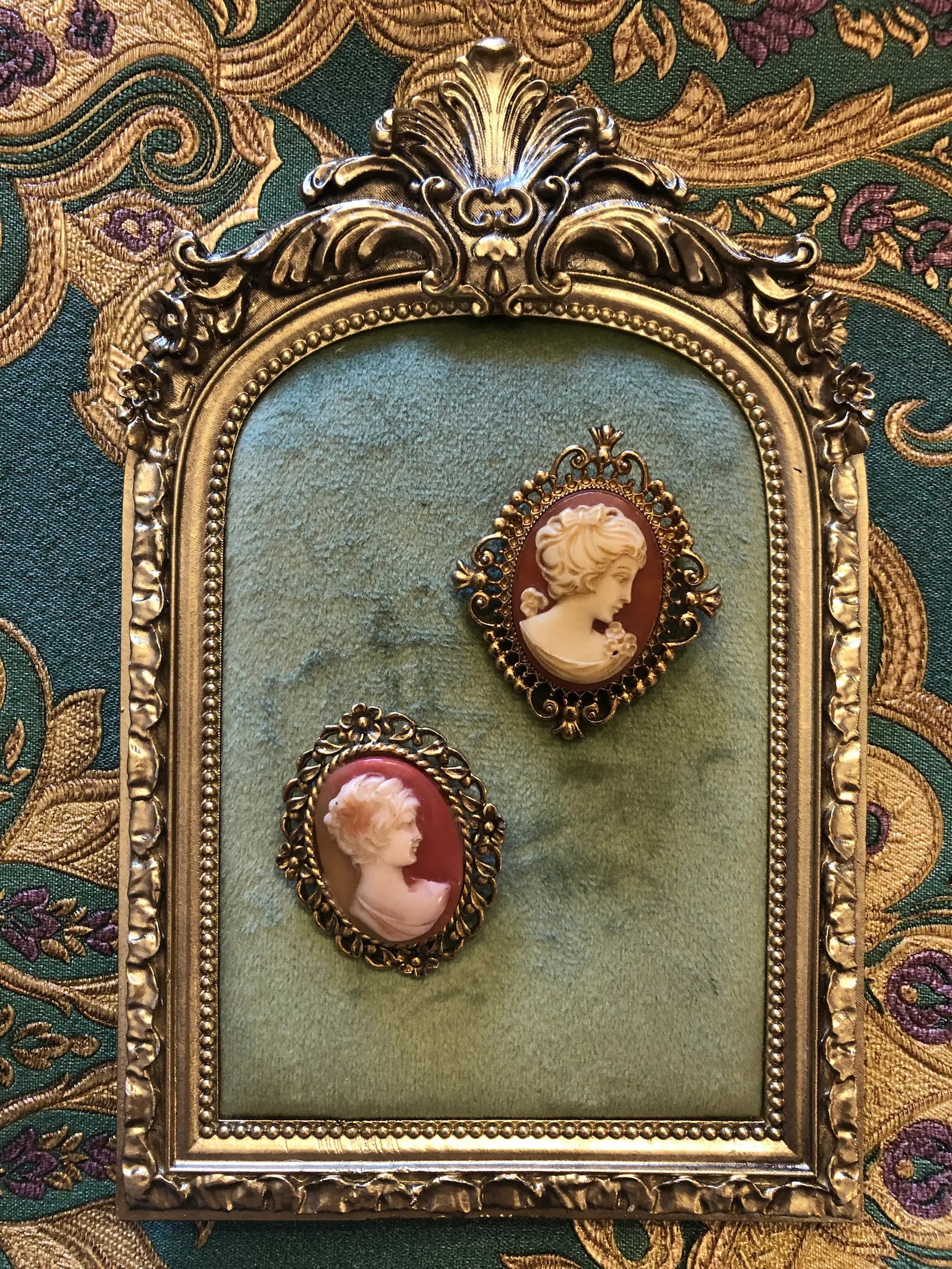 An ornate vintage-style framed brooch display featuring two  Broochella oval cameo brooches  with pink and cream profiles, mounted on a green velvet backing inside a carved gold frame, set against a patterned green and gold fabric background.