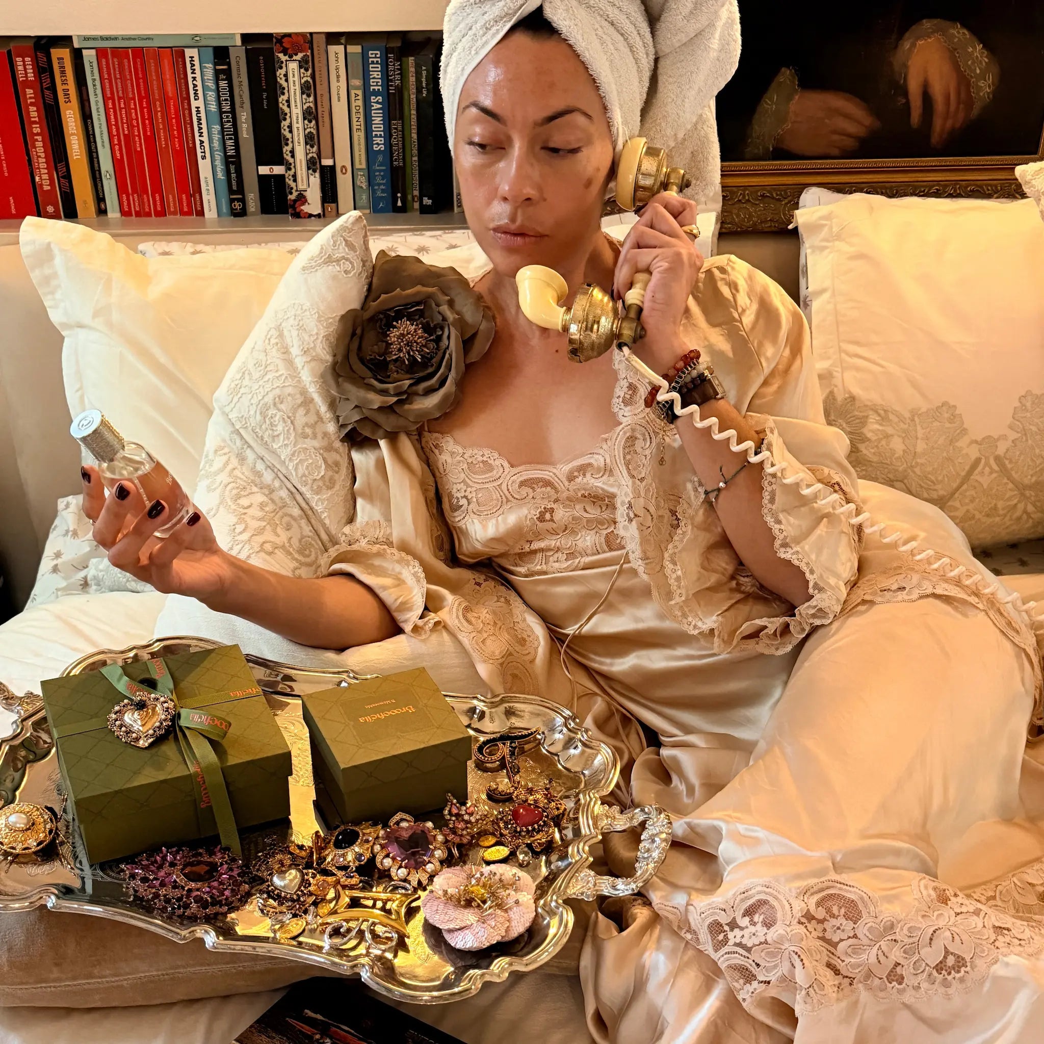 A woman in a beige lace robe and white towel wrap sits on a sofa holding a vintage-style gold telephone, with a tray of Broochella and green gift boxes in the foreground. She is holding a bottle of the Broochella Eau du Parfum.