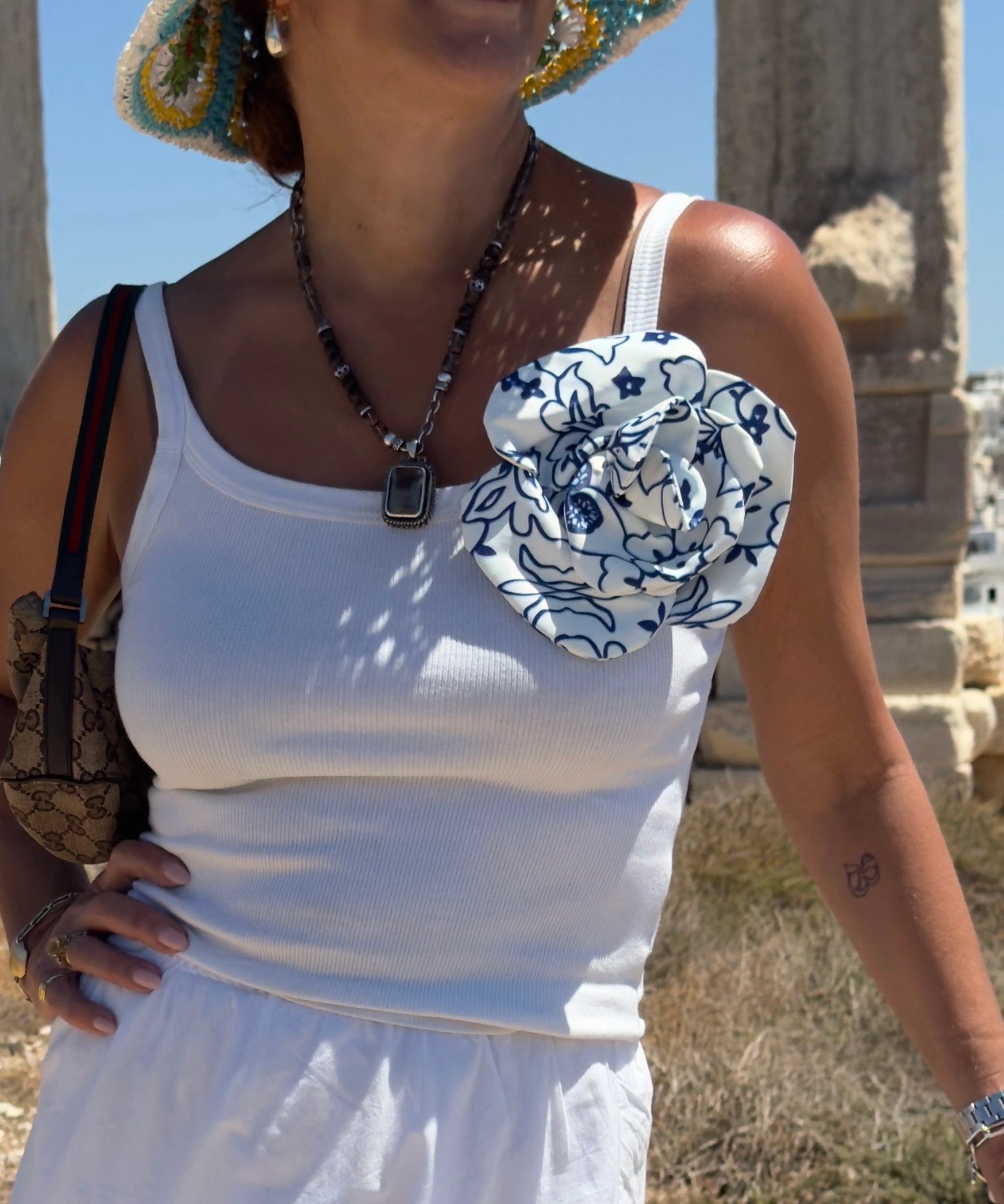 A woman outdoors wearing a white ribbed tank top with a  Broochella Electra fabric flower brooch  featuring a blue floral pattern on the chest. She is also wearing a patterned hat, a beaded necklace, and a patterned shoulder bag.