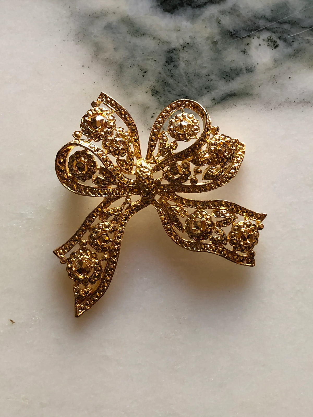 Broochella Elisabetta Vintage Gold Bow Brooch featuring an ornate sculptural ribbon design with floral and scroll openwork detailing on a cream and grey marble background.