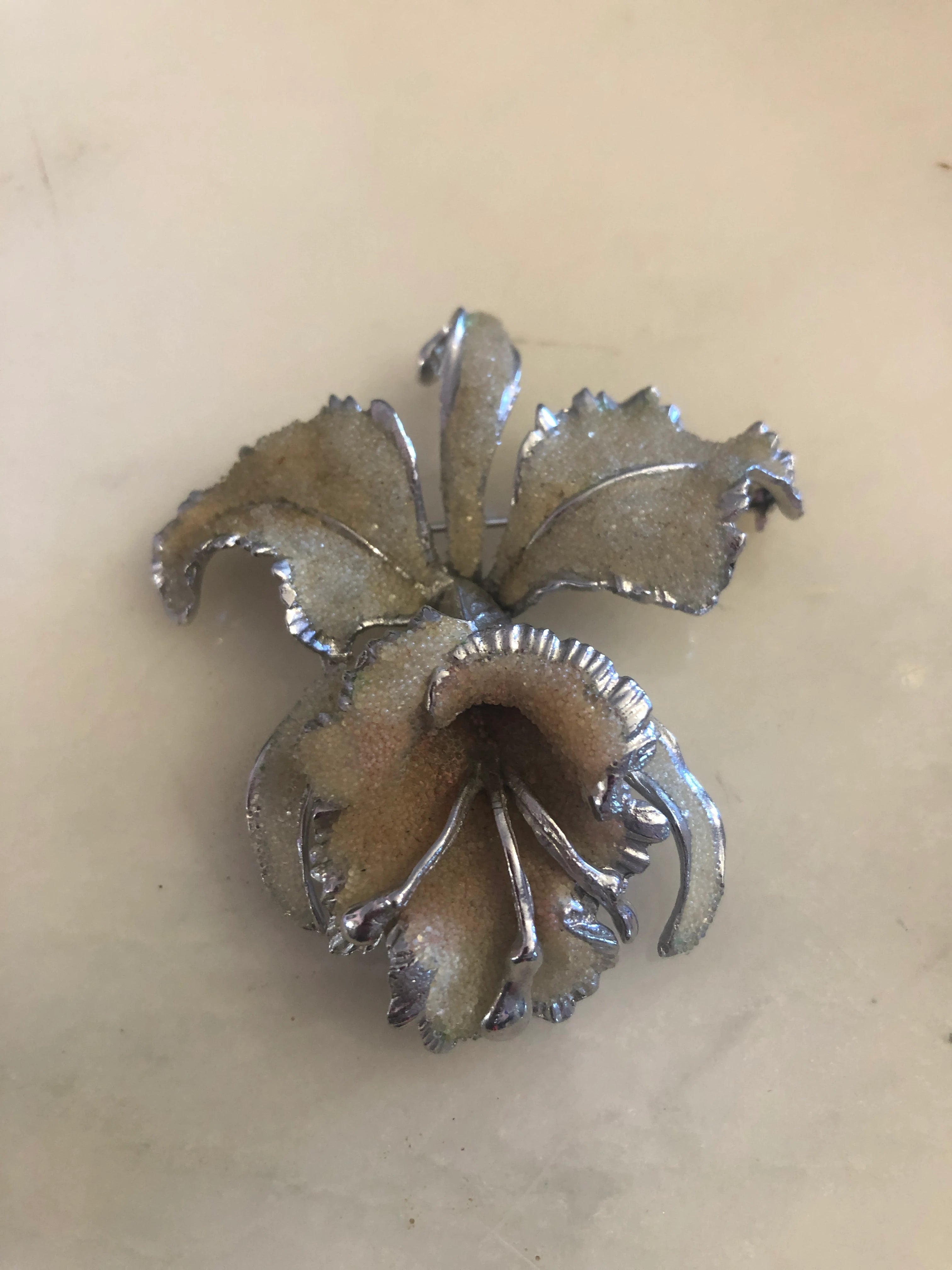 A vintage signed Elizabeth Reimer Arcansas Coralene silver-tone orchid brooch featuring five sculptural petals with a fine frosted sand-texture coralene surface, polished serrated silver-tone metal petal edges and a three-dimensional trumpet centre. The background is white marble