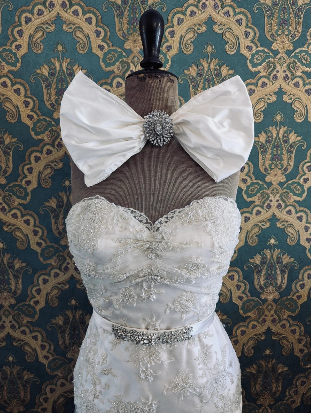 Broochella large handmade ivory bridal bow, with a crystal and silver tone metal centre embellishment. Pinned on a mannequin that is wearing a white, beaded strapless wedding dress. The background is green and gold damask wallpaper. 