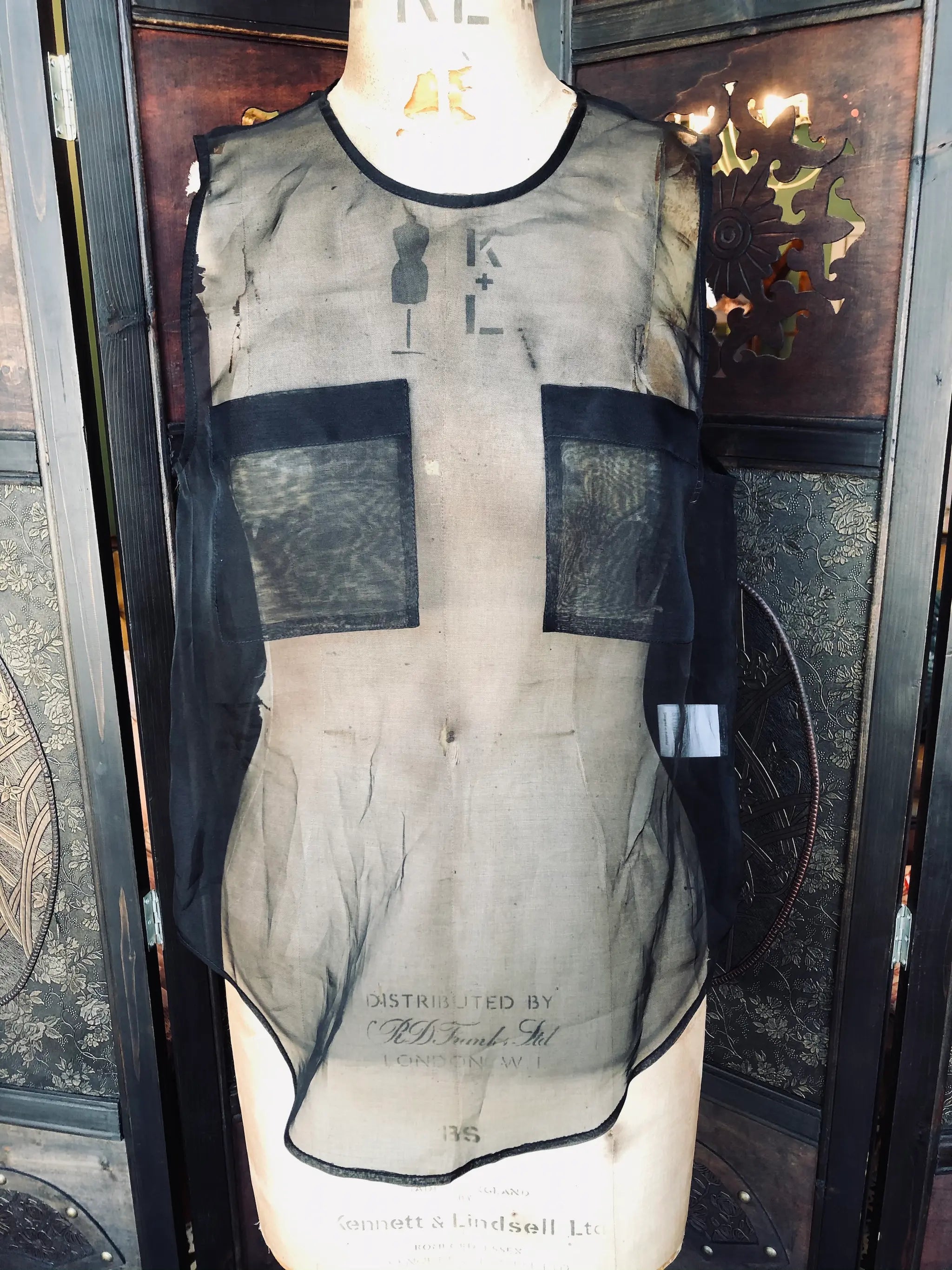 Broochella Emerson studios, Small, 100% Silk, Black, Sleeveless design, Sheer silk finish, Front panel pockets. Keyhole back detail. It is presented on a vintage mannequin. The background is a ornate, vintage, dark wood room divider. 