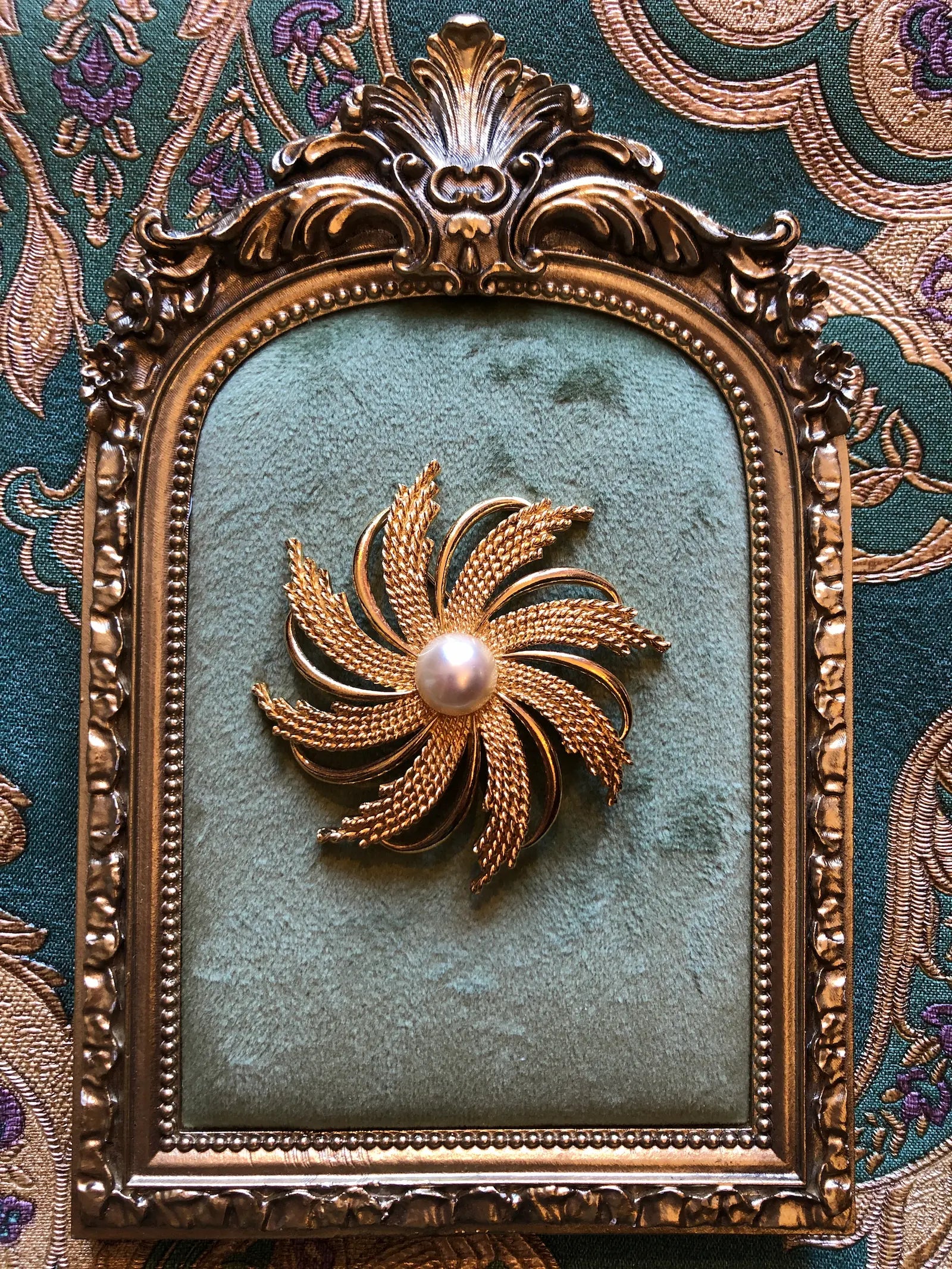 A Broochella Emma Page vintage sunburst gold-tone brooch with a central pearl, mounted on a square teal velvet panel within an ornate gold frame, set against a patterned teal and gold fabric background.