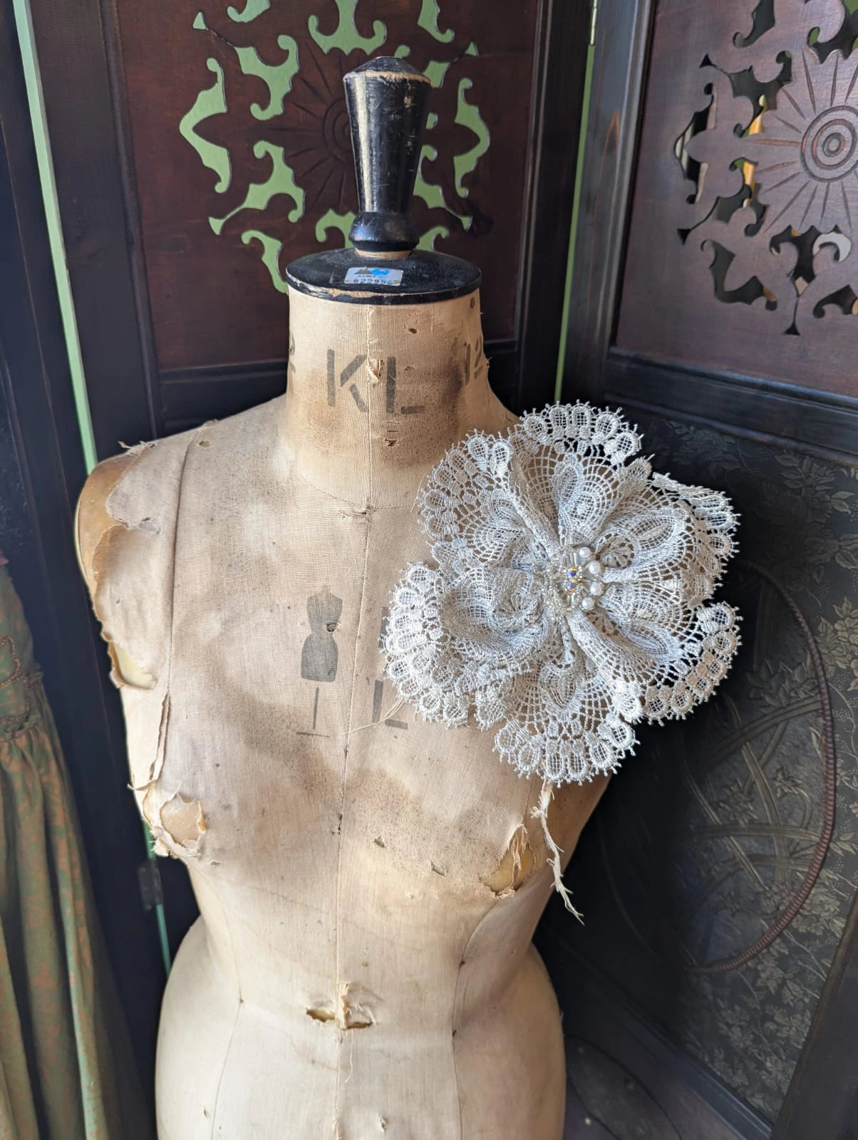 Broochella etoile de dentelle ivory lace handamde fabric flower brooch with beaded centre. It is pinned to an old mannequin. The background is a decorative brown wooden screen.