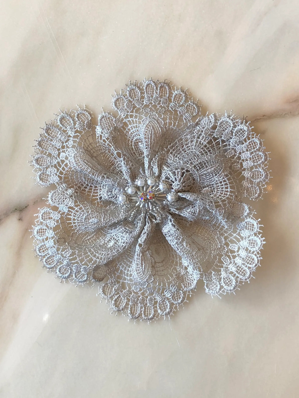 Broochella Étoile de Dentelle handmade designer lace flower brooch in shimmering silver-grey lace, with sculptural layered petals, pearl accents and a crystal centre, photographed on pale cream marble with natural grey veining.