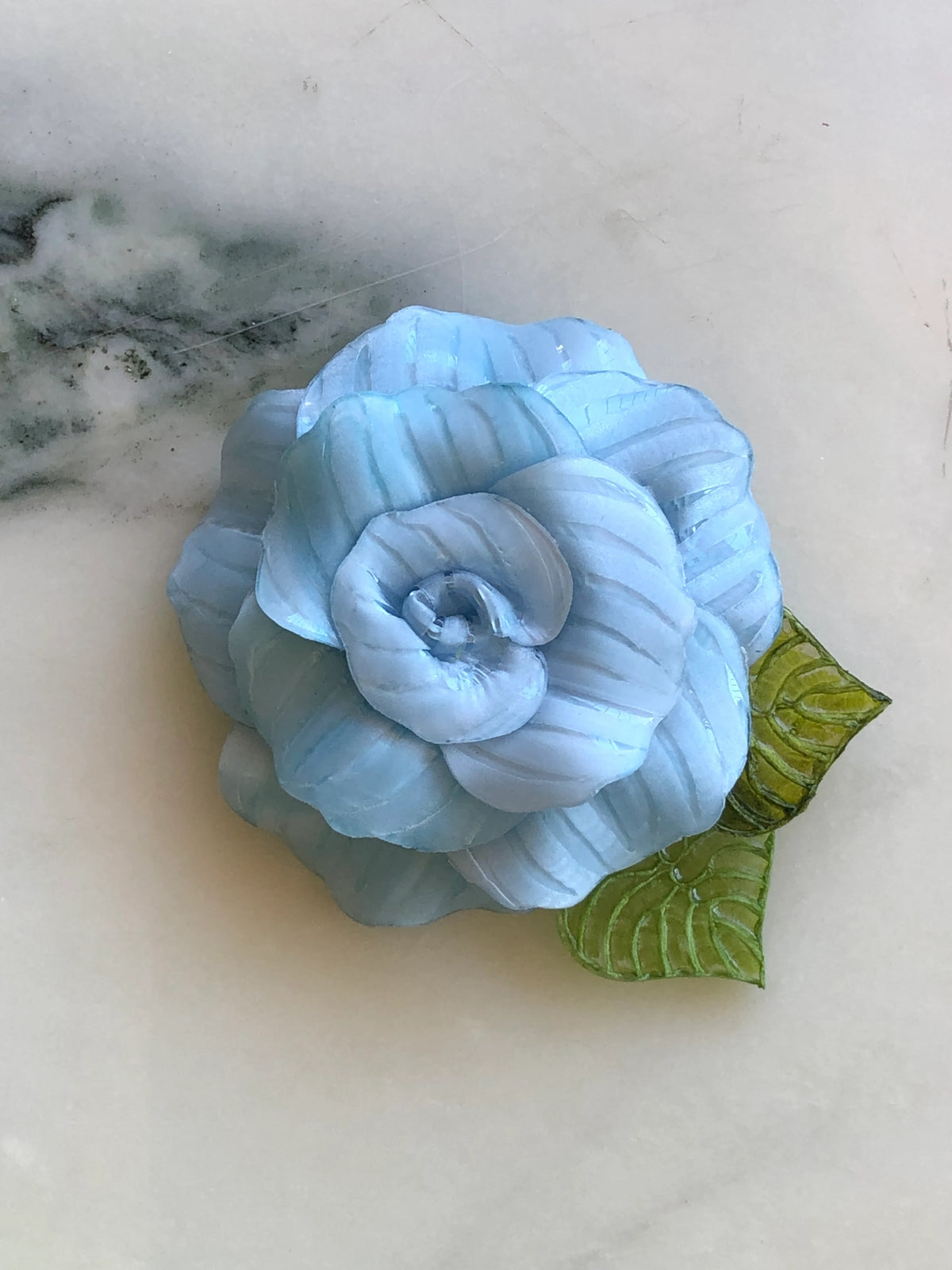 Broochella Fabrice Paris Camelia flower brooch for men in pale blue striped fabric, featuring layered sculptural petals and two textured green leaves. A contemporary men's floral lapel accessory photographed on white marble with grey-green veining.