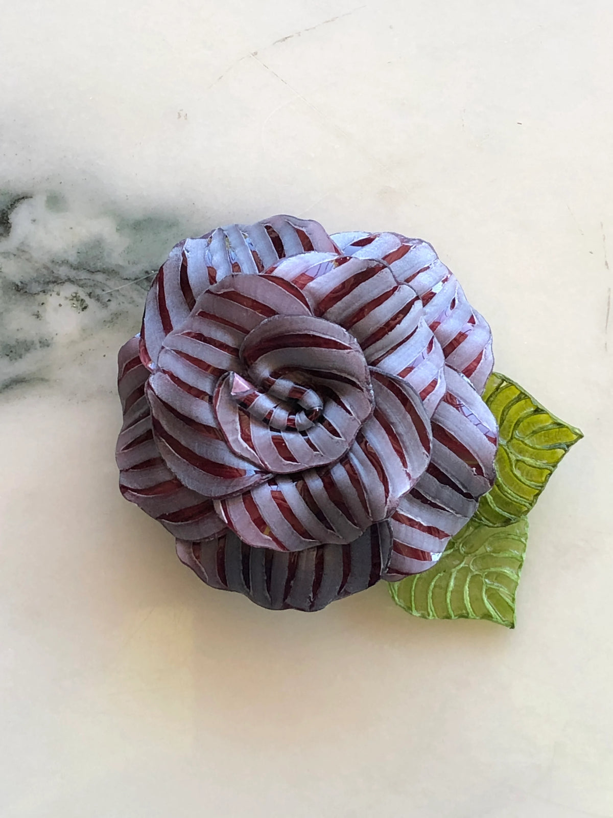 Broochella Fabrice Paris Camelia flower brooch for men in burgundy and pale mauve striped fabric, featuring layered sculptural petals and two textured green leaves. An expressive men's floral lapel accessory photographed on white marble with grey-green veining.