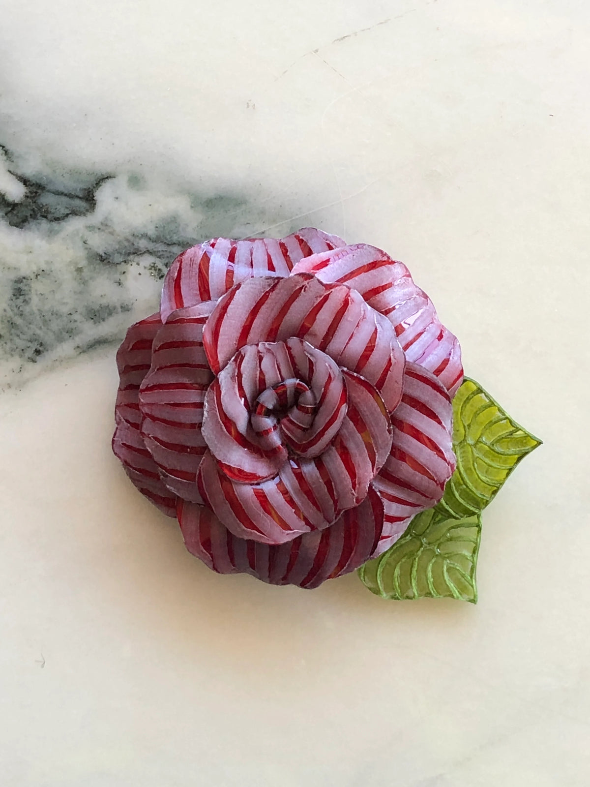 Broochella Fabrice Paris Camelia flower brooch for men in red and soft pink striped fabric, featuring layered sculptural petals and two textured green leaves. A statement men's floral lapel accessory photographed on white marble with grey-green veining.