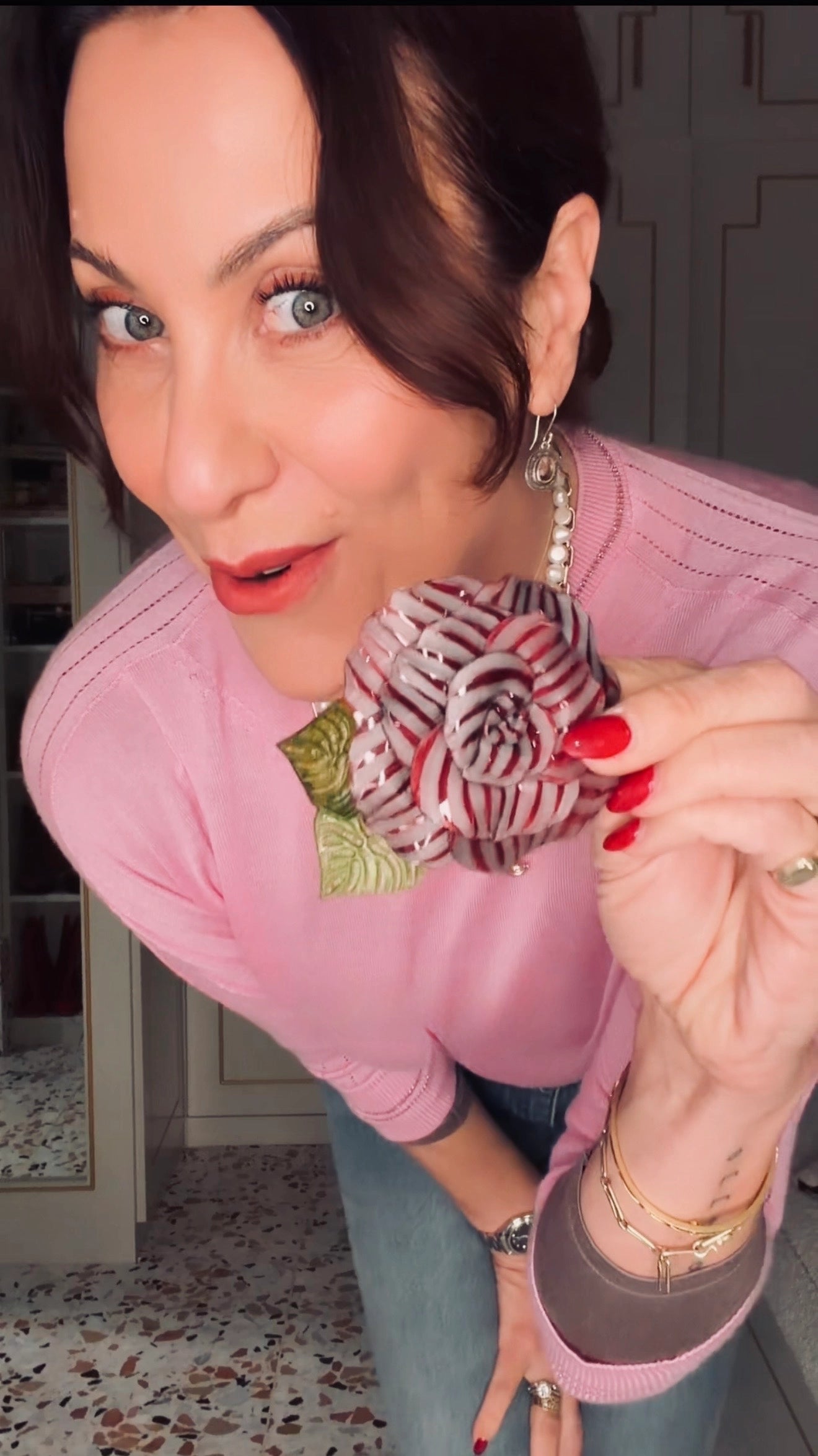 A woman in a pink sweater holds up a Broochella x Fabrice Camelia flower brooch with red and white stripes and green leaves, showing it close up in an indoor setting.