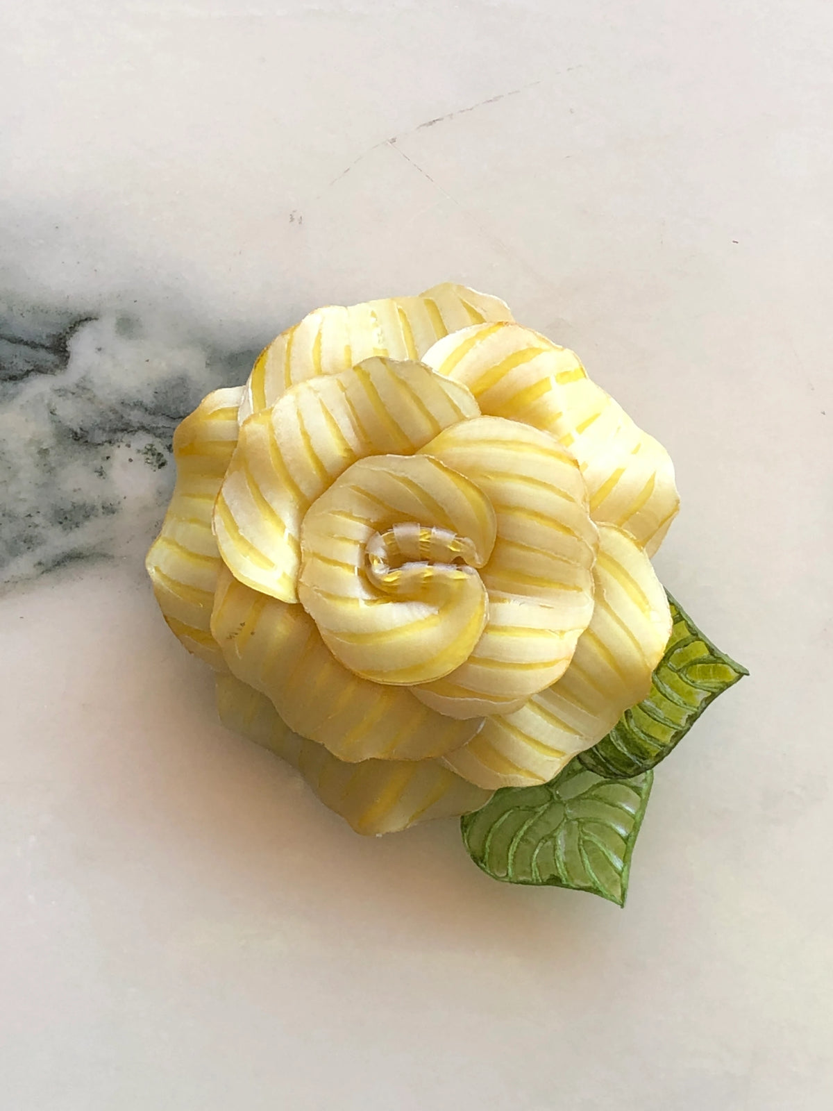 Broochella Fabrice Paris Camelia flower brooch for men in yellow and ivory striped fabric, featuring layered sculptural petals and two textured green leaves. A bold men's floral lapel accessory photographed on white marble with grey-green veining.