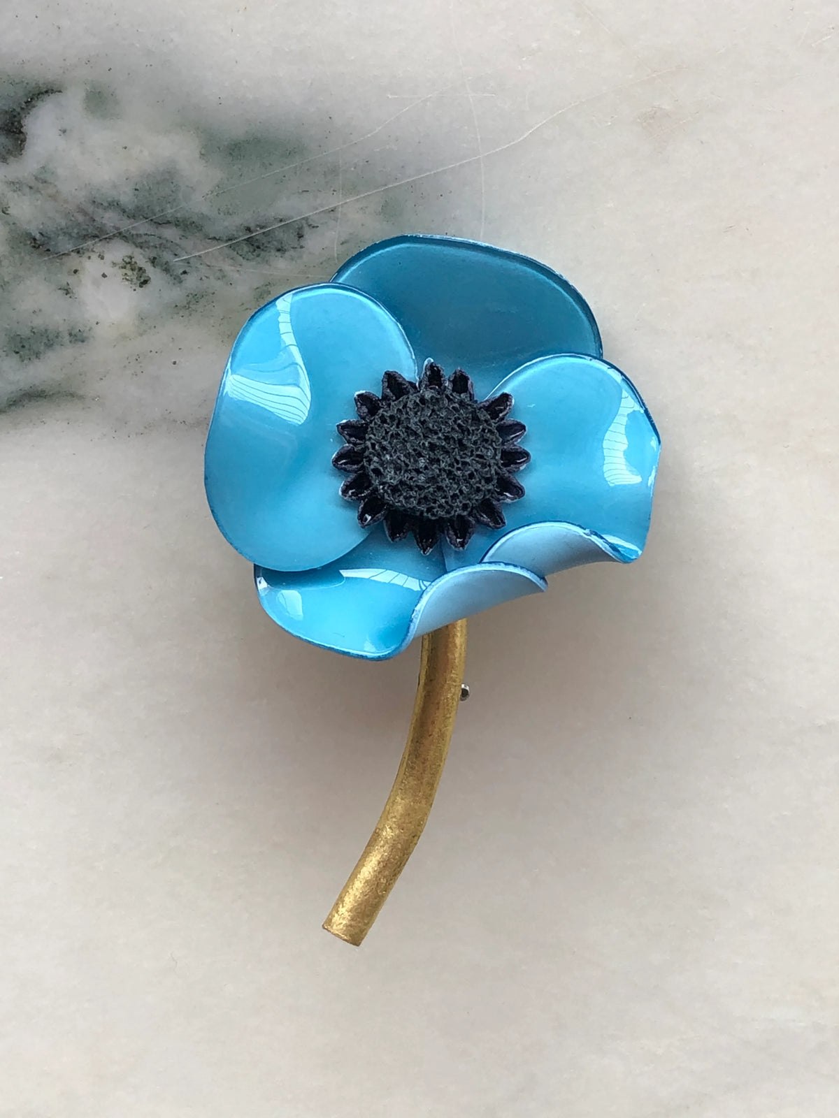 Broochella Fabrice Paris Poppy Flower Brooch in vibrant turquoise blue with glossy layered petals, a textured black centre and curved antique gold stem. Photographed on white marble with grey-green veining.