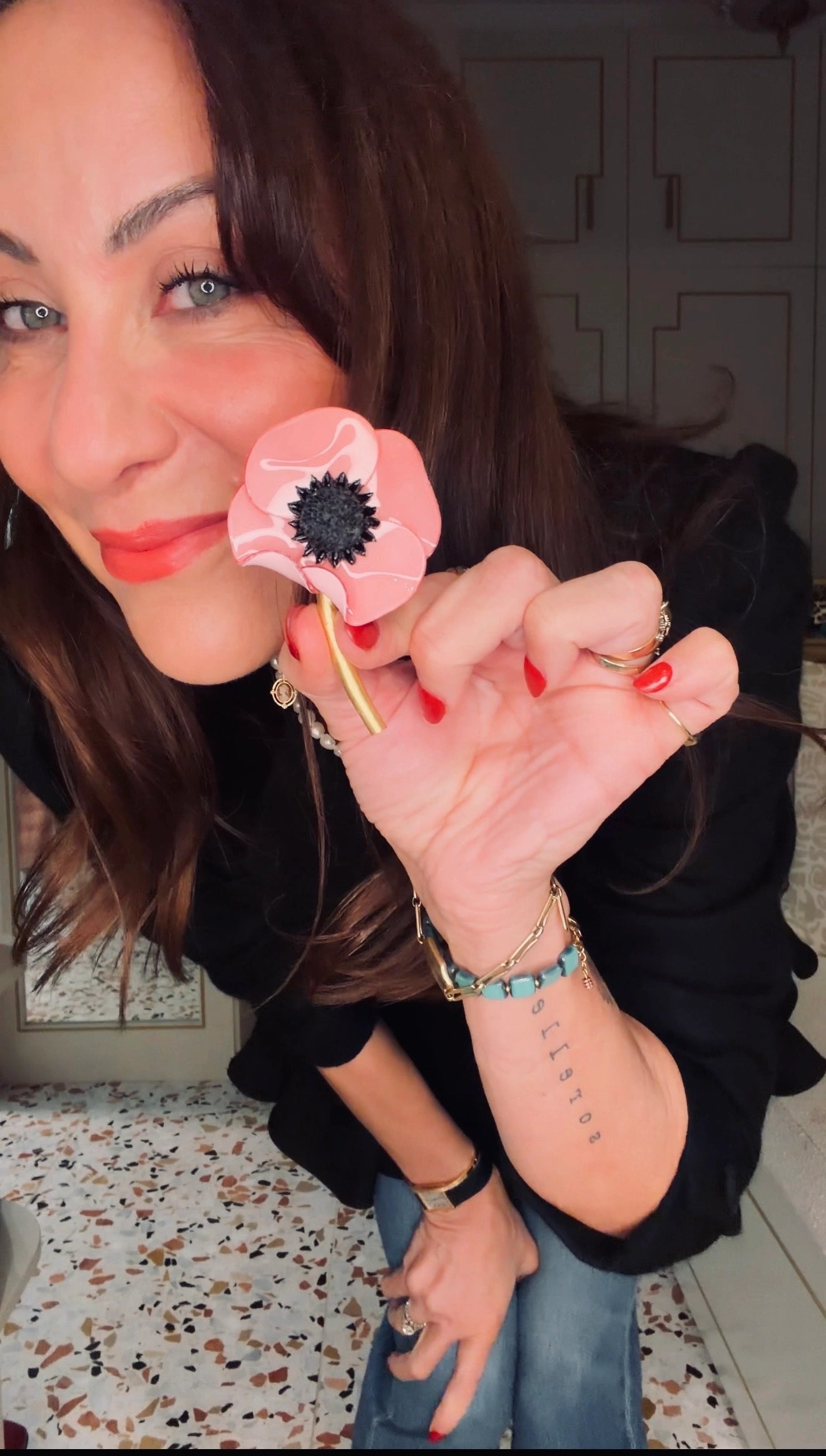 A woman holding a Broochella x Fabrice pink acetate poppy flower brooch  with a black centre, wearing a black top and blue jeans in an indoor setting.