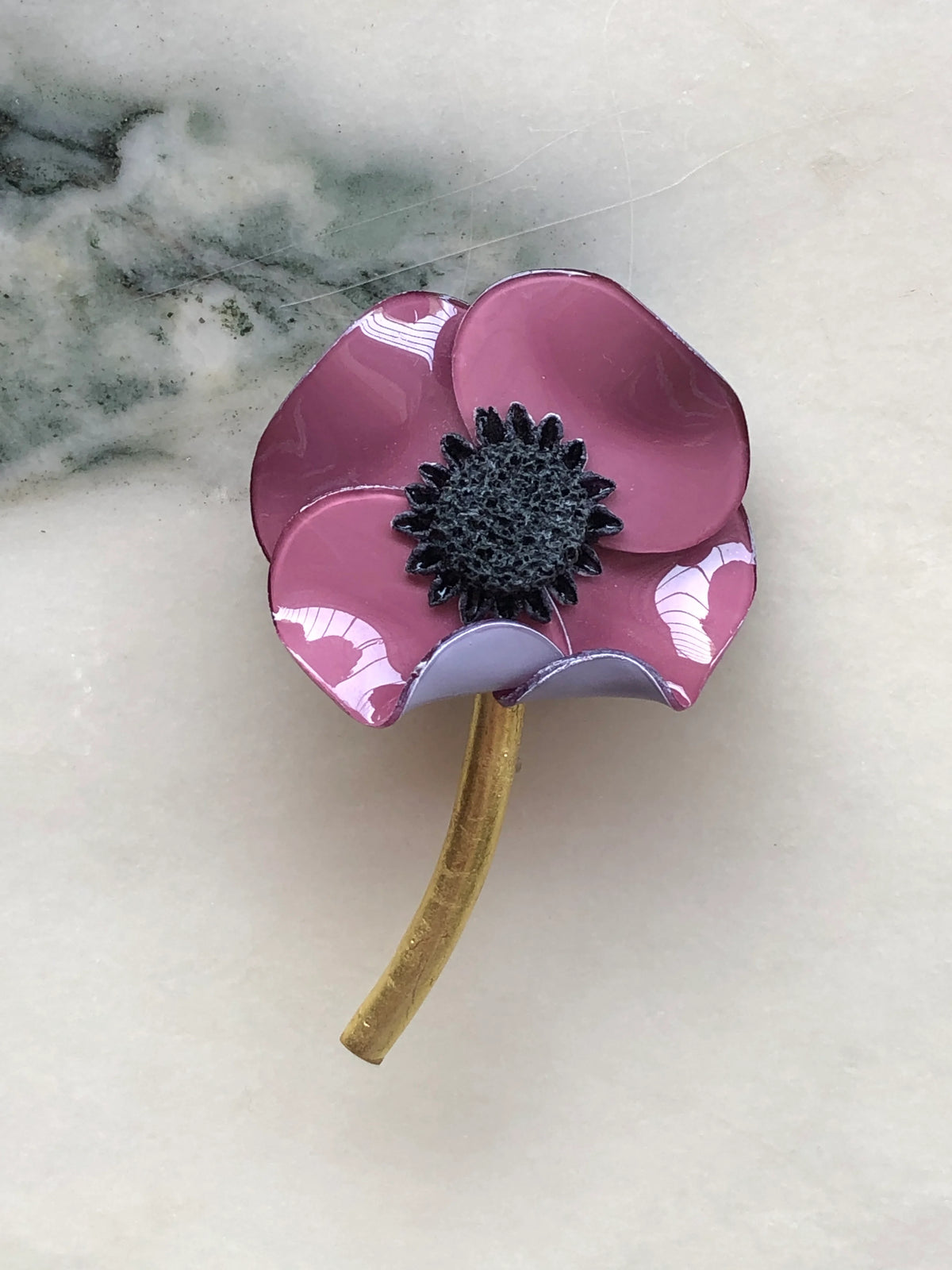 Broochella Fabrice Paris Poppy Flower Brooch for men in purple, featuring glossy sculptural petals, a textured black centre and curved antique gold stem. A statement floral lapel pin for men's jackets, blazers and suits, photographed on white marble with grey-green veining.