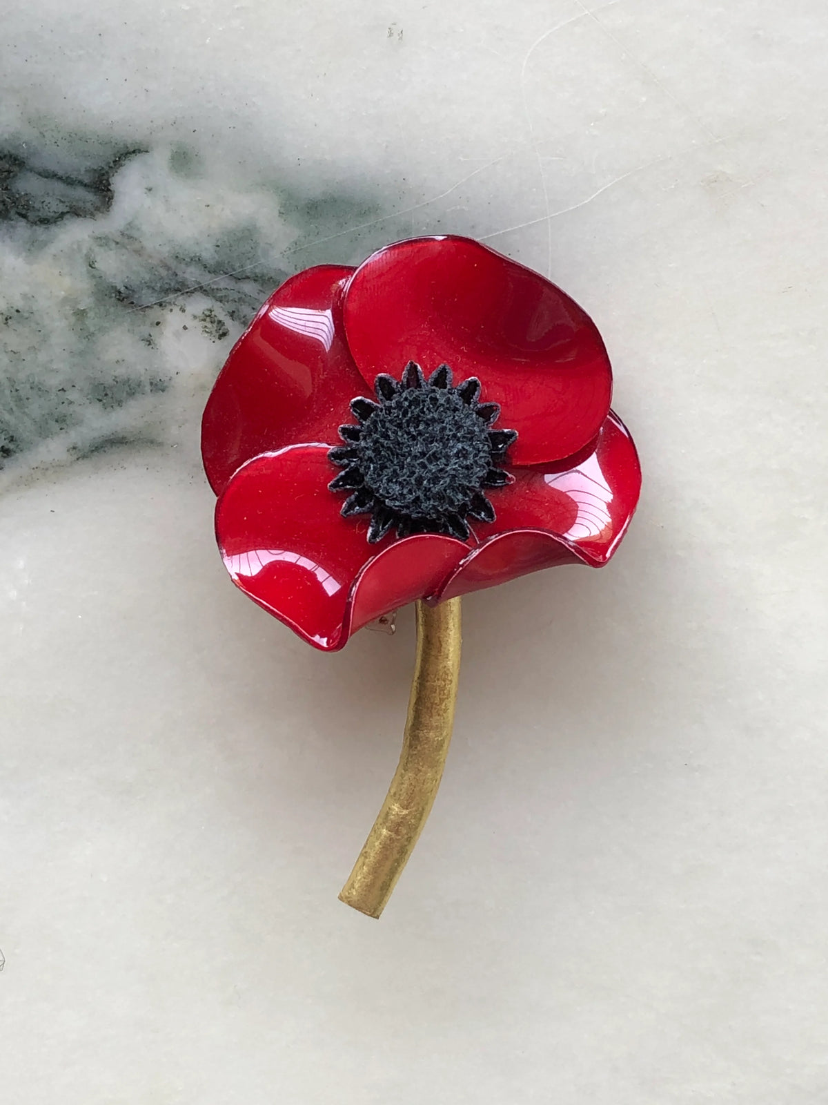 Broochella Fabrice Paris Poppy Flower Brooch in vibrant red with glossy sculptural petals, a textured black centre and curved antique gold stem. Photographed on white marble with grey-green veining.