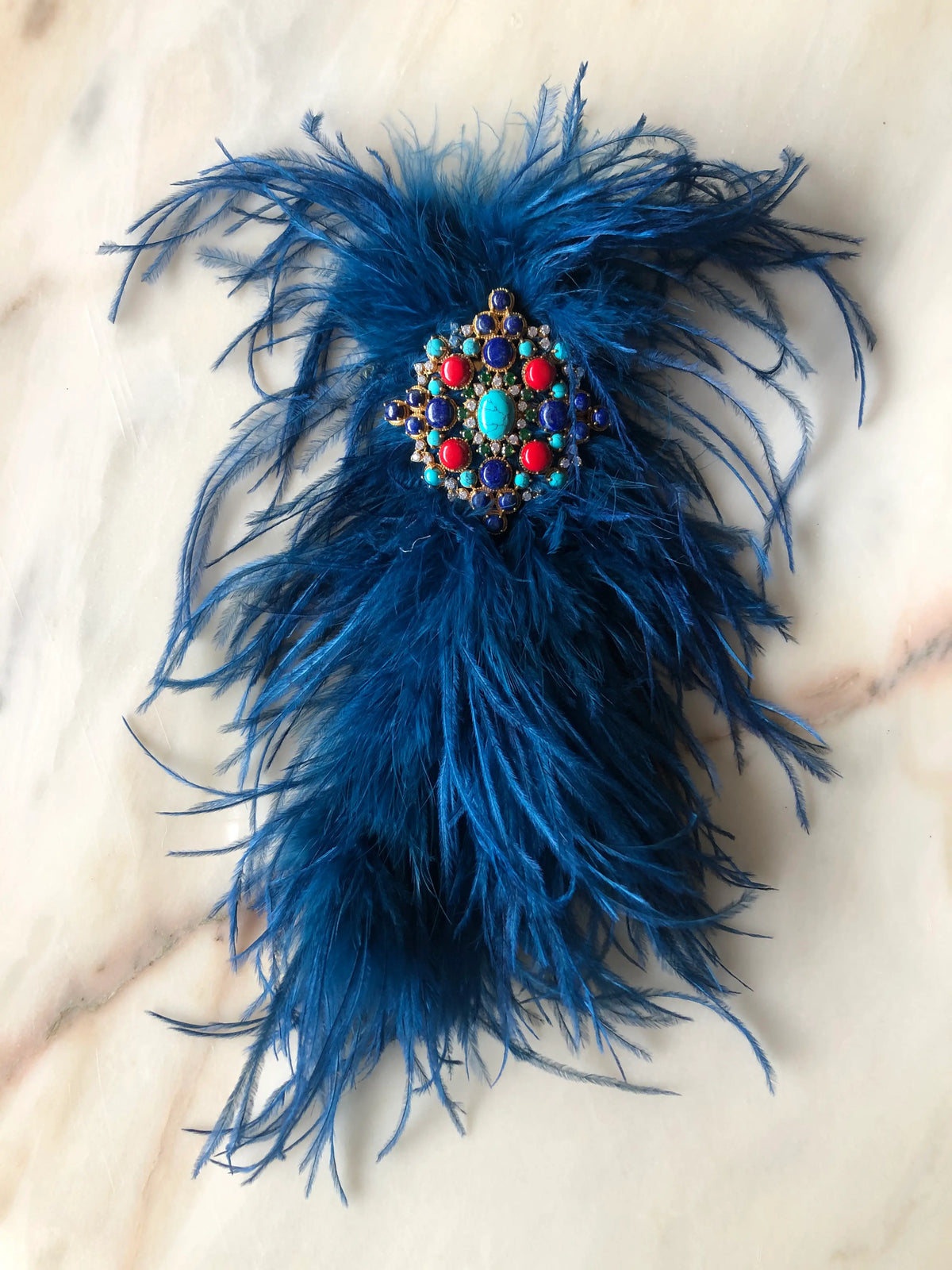Broochella blue Feather Bejewelled Brooch with cascading ostrich feathers and an ornate turquoise, red and blue jewelled centre, photographed on a white marble background.