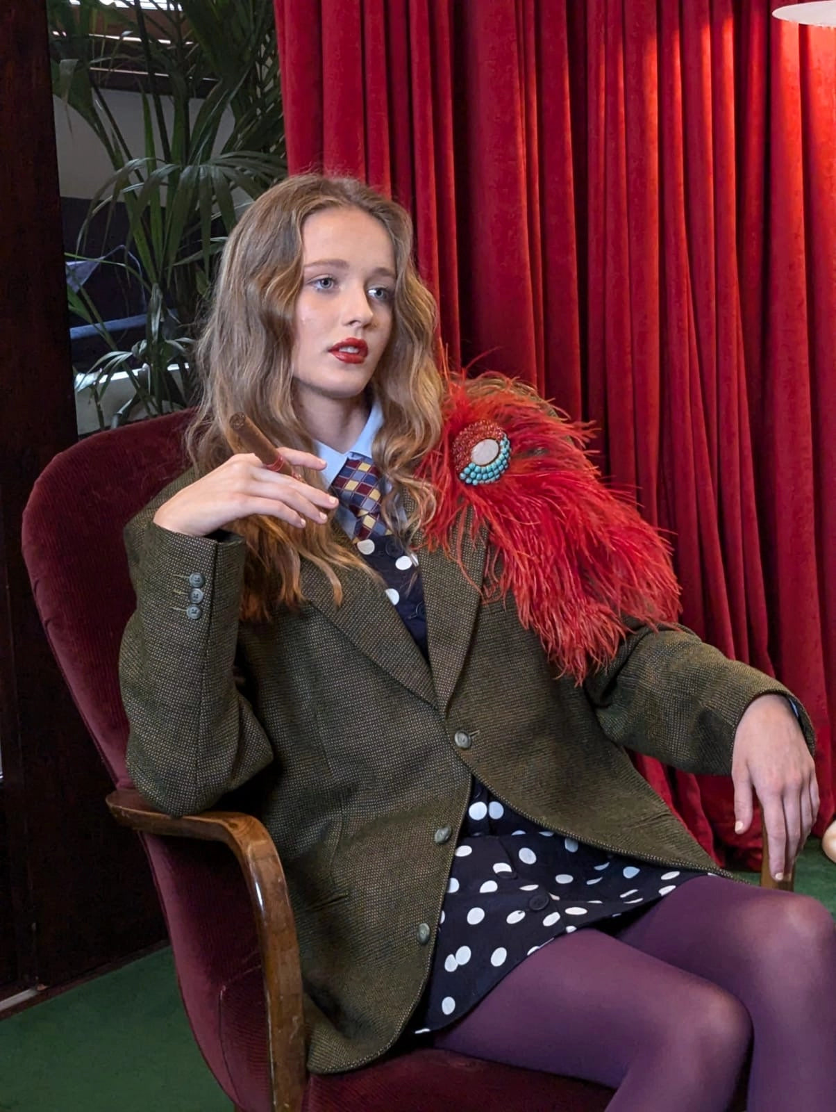 A woman seated in a red velvet armchair wearing an olive green tweed blazer, a black dress with white polka dots, a patterned tie, and purple tights. She has a broochella bejewelled  feathered brooch accessory and a decorative brooch on her lapel, set against a backdrop of red curtains.