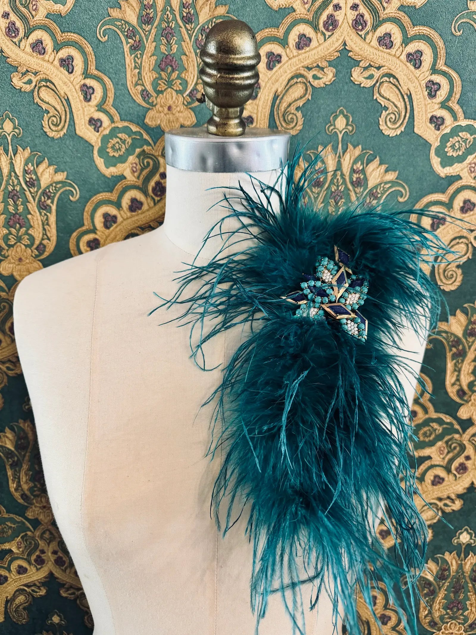 Broochella teal handmade large feather brooch with statement brooch centre embellishment.  Pictured on a mannequin.  The background is green and gold damask wallpaper.