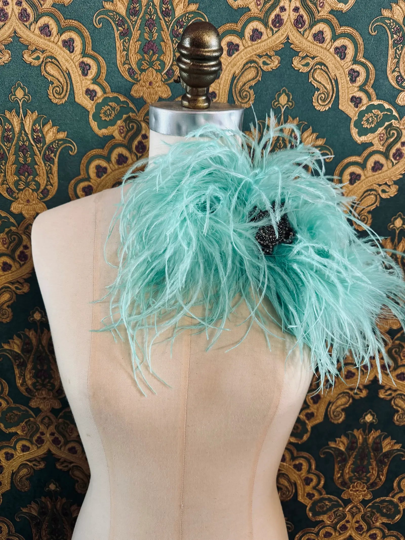 Broochella aqua handmade large feather bow brooch with embellished centre.  Pictured on a mannequin.  The background is green and gold damask wallpaper.