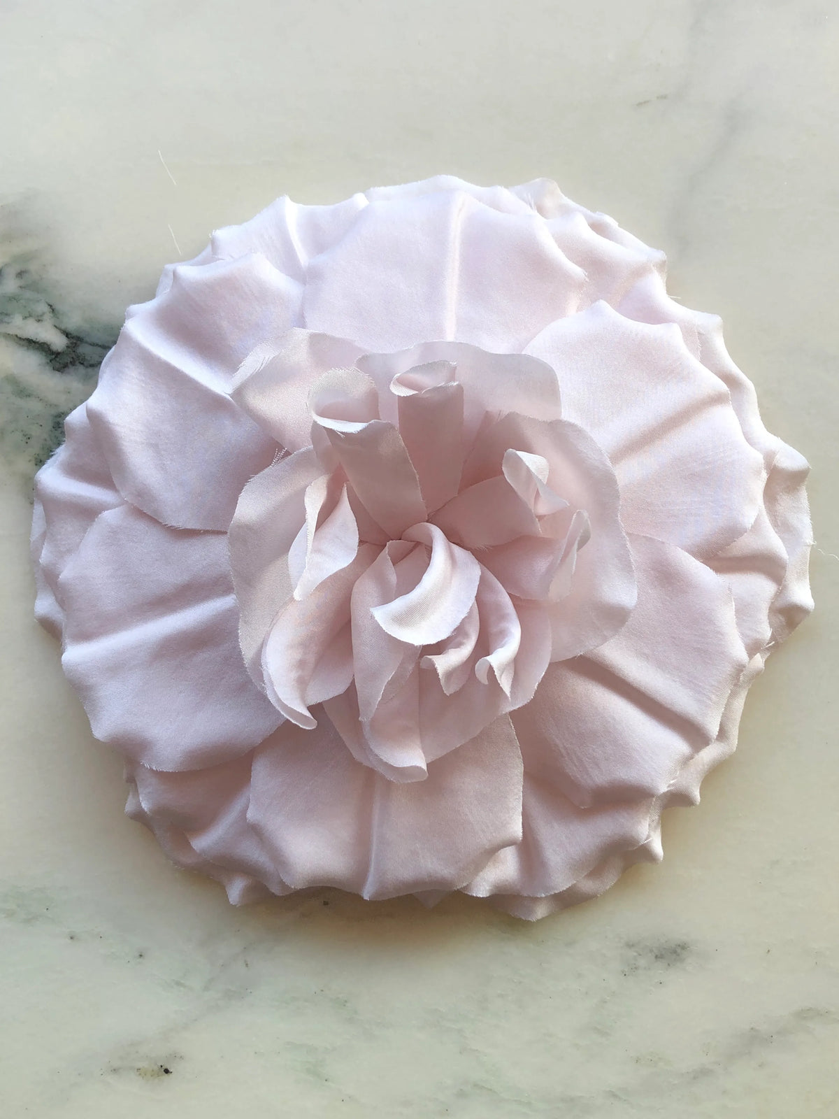 Broochella Femmina Flower Brooch Pin in soft blush pink with oversized layered fabric petals and a sculptural ruffled centre, photographed on a white marble background with grey veining.