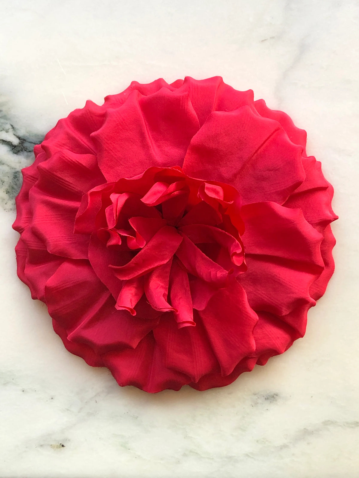 Broochella Femmina Flower Brooch Pin in vibrant red with oversized layered fabric petals and a sculptural ruffled centre, photographed on a white marble background with grey veining.