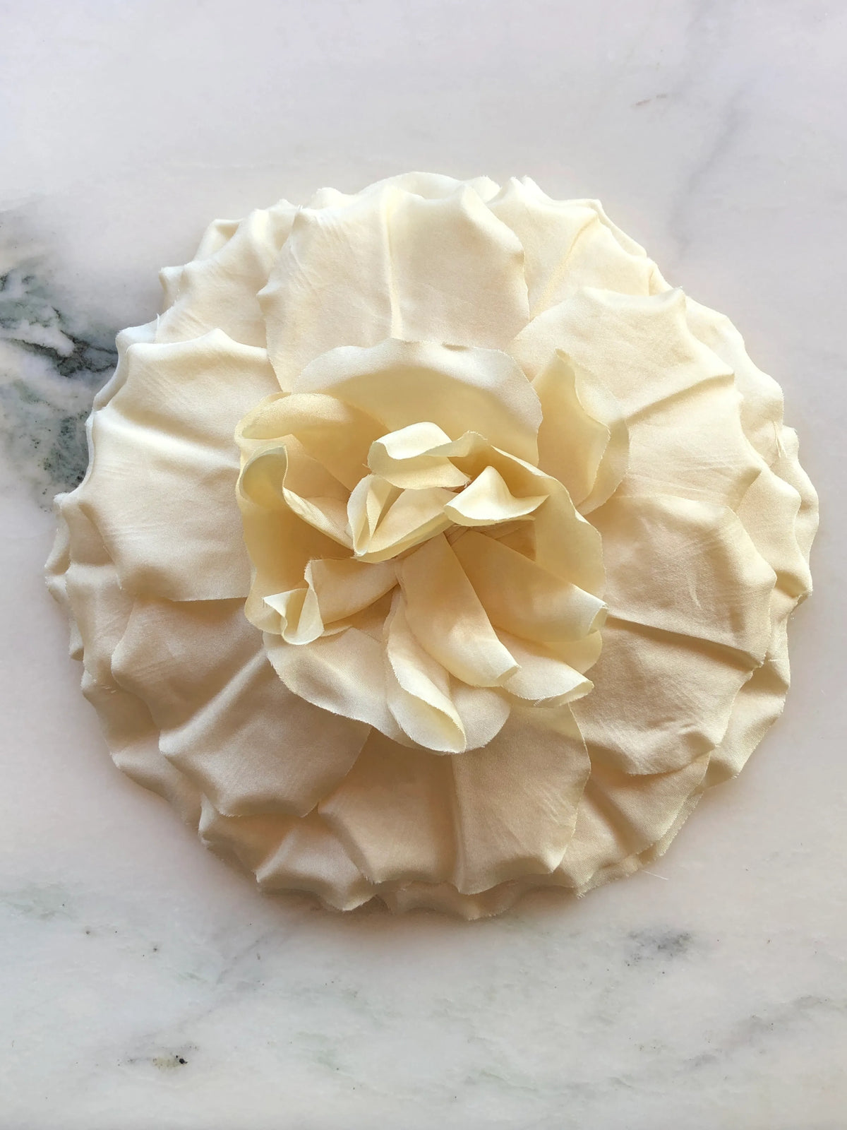 Broochella Femmina Flower Brooch Pin in soft buttery yellow with oversized layered fabric petals and a sculptural ruffled centre, photographed on a white marble background with grey veining.