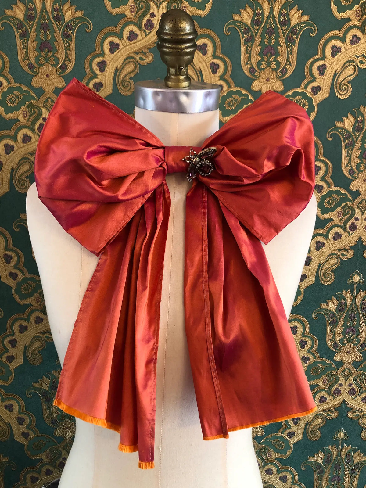 Broochella Fiocco Bejewelled Fashion Bow Brooch in orange silk with an ornate jewelled insect centre and long flowing tails, styled on a cream mannequin against ornate green and gold wallpaper.