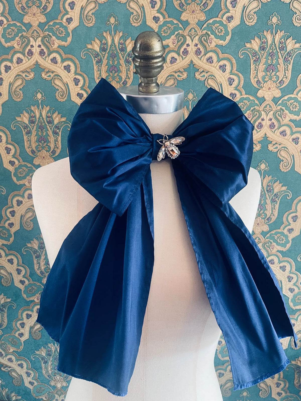 Broochella Fiocco large handmade silk embellished bow in blue. Pictured on a mannequin. The background is green and gold damask wallpaper.