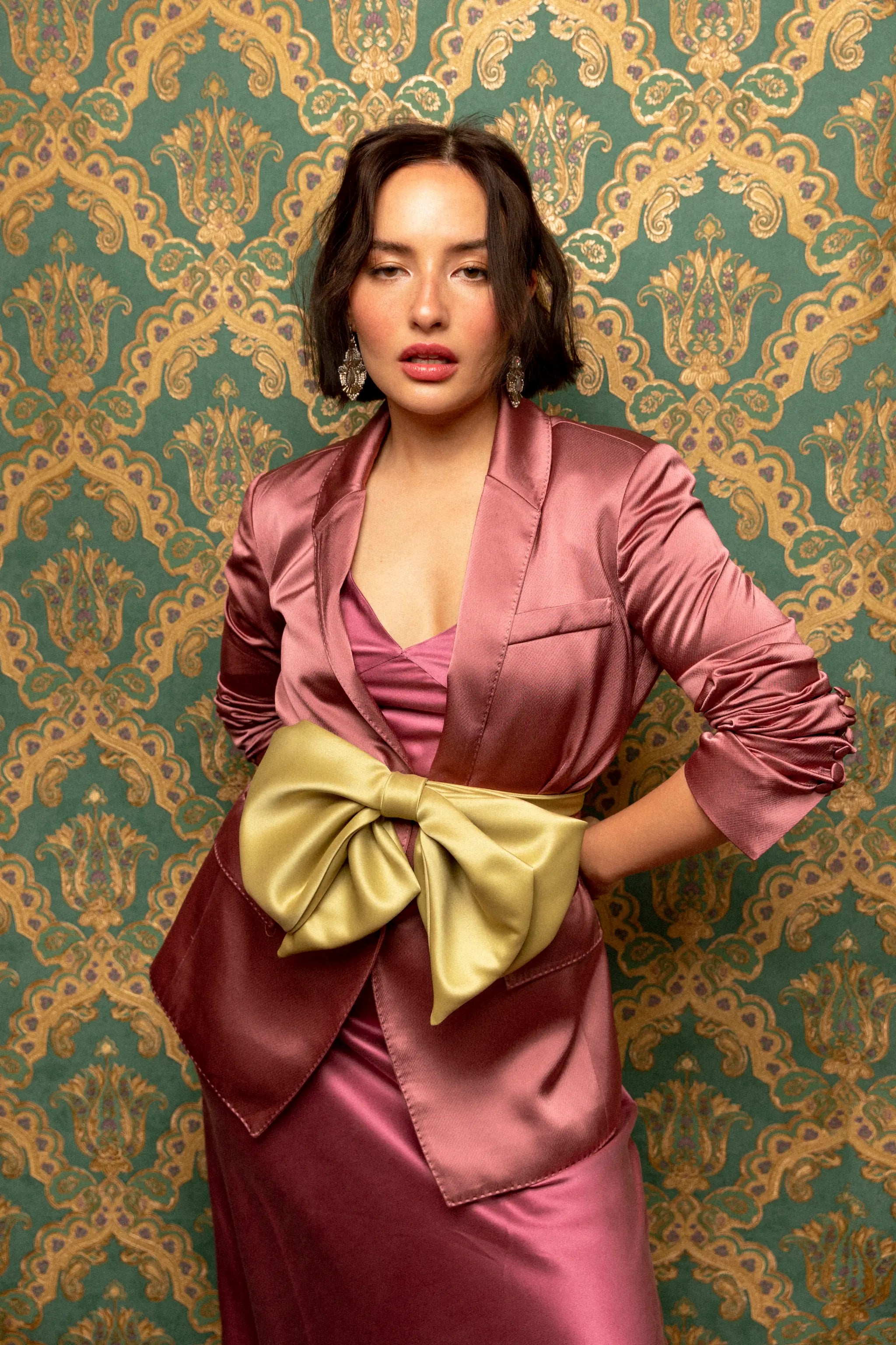 Broochella Fiocco small green handmade  satin bow brooch. Pictured on a female model in a pink dress and jacket. The background is green and gold damask wallpaper.