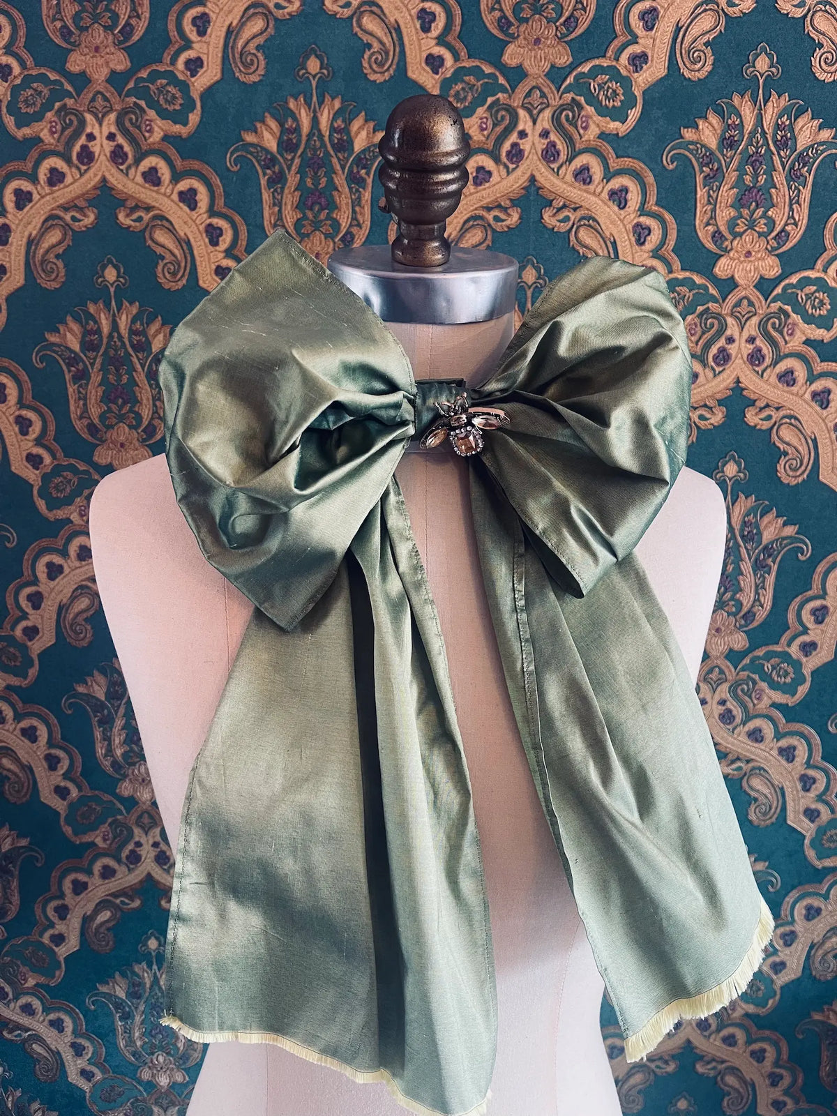 Broochella Fiocco large handmade silk embellished bow in mint. Pictured on a mannequin. The background is green and gold damask wallpaper.
