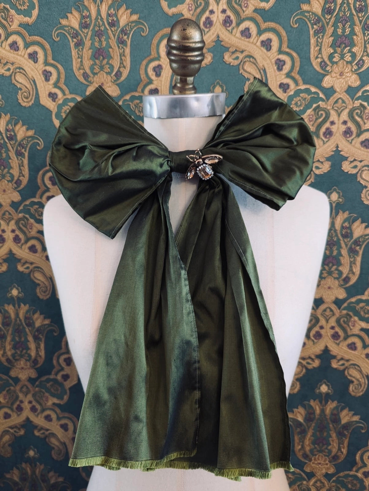 Broochella Fiocco large handmade silk embellished bow in green. Pictured on a mannequin. The background is green and gold damask wallpaper.
