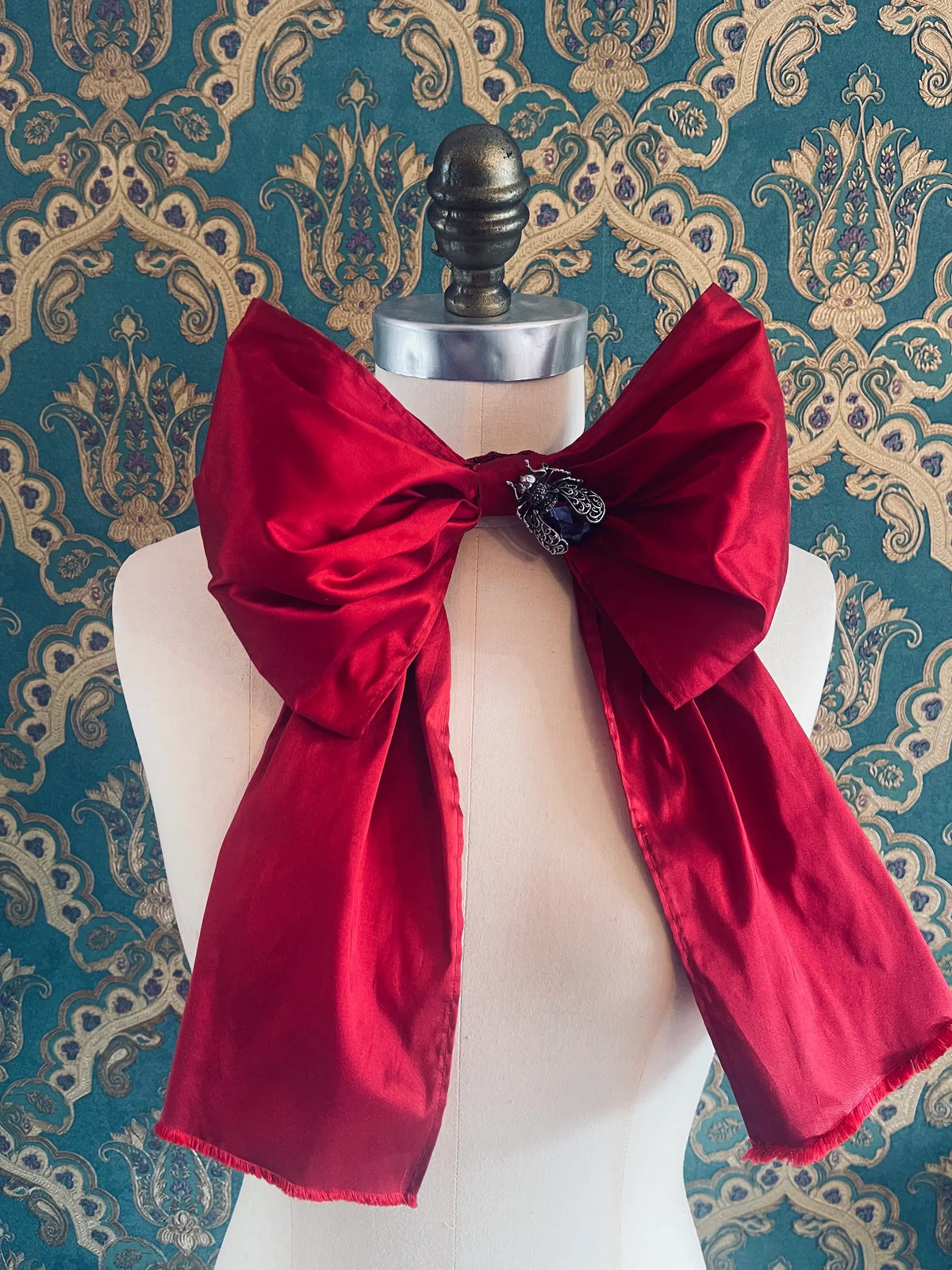 Broochella Fiocco large handmade silk embellished bow in red. Pictured on a mannequin. The background is green and gold damask wallpaper.