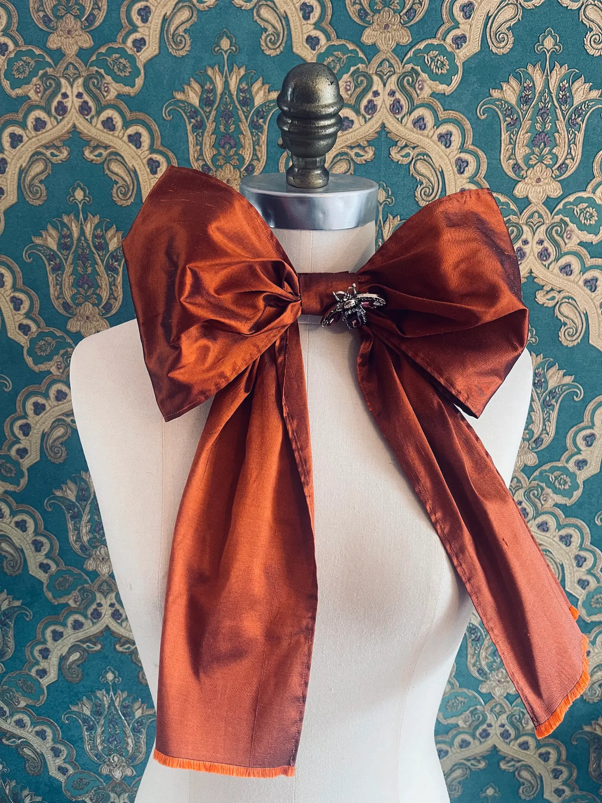Broochella Fiocco large handmade silk embellished bow in rust. Pictured on a mannequin. The background is green and gold damask wallpaper.