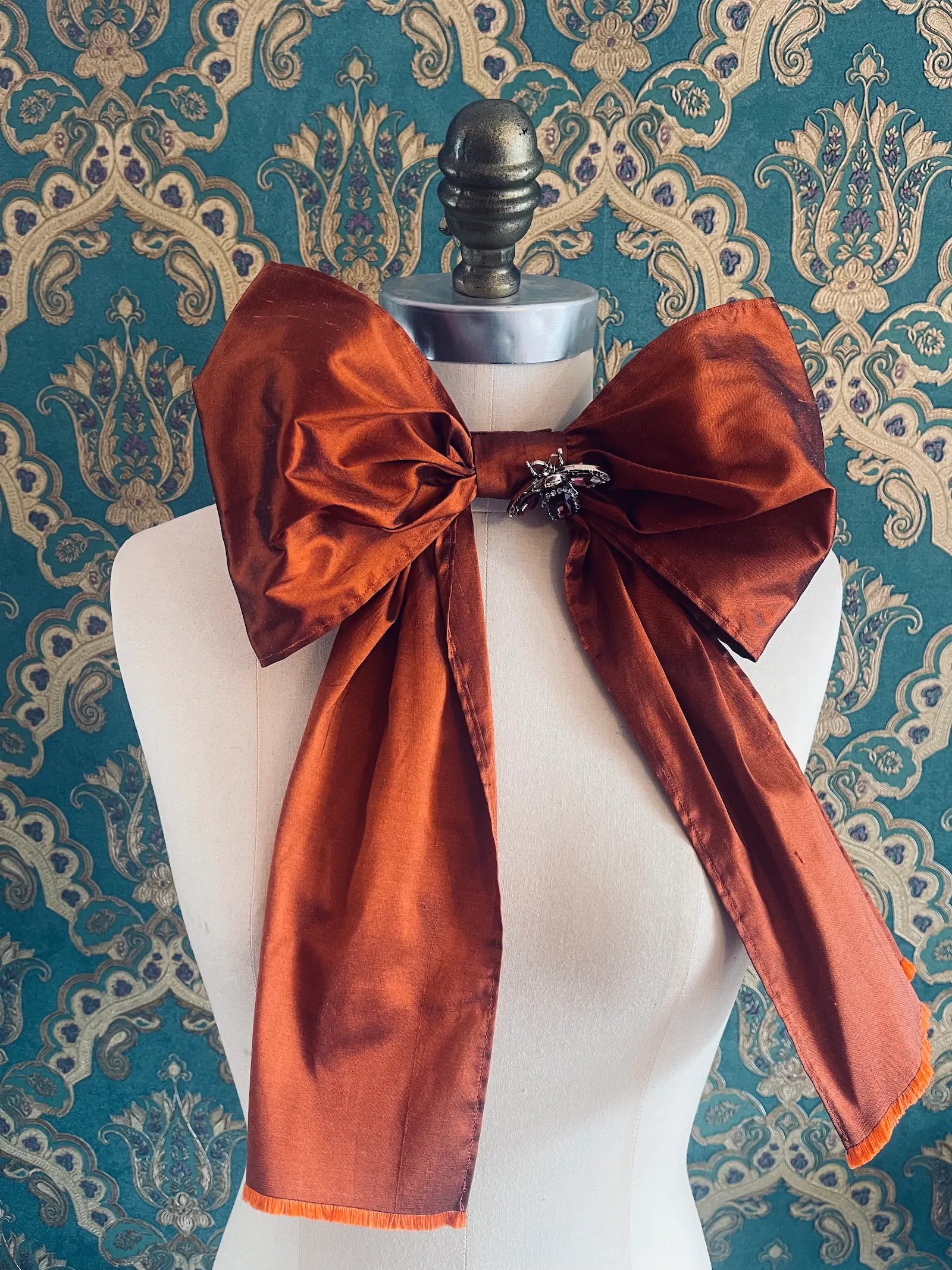 Broochella Fiocco large handmade silk embellished bow in rust. Pictured on a mannequin. The background is green and gold damask wallpaper.