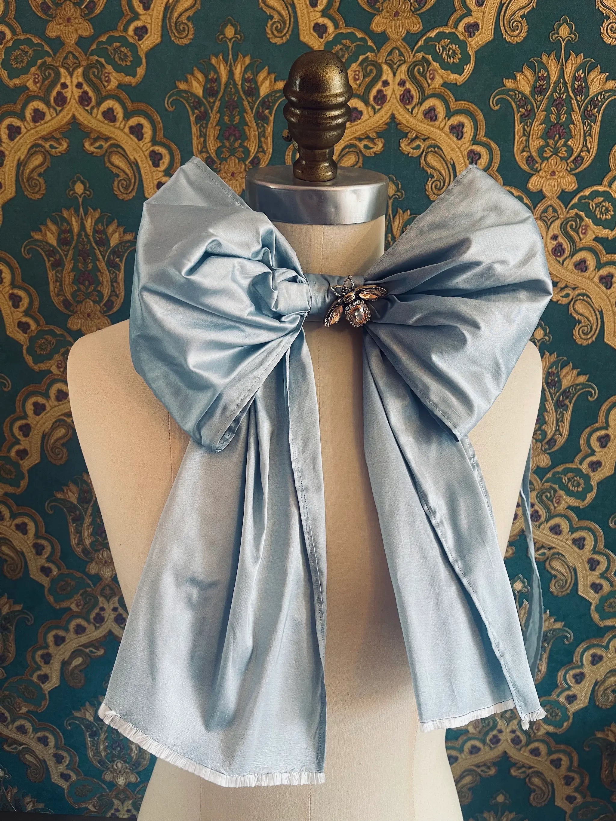 Broochella Fiocco large handmade silk embellished bow in sky blue. Pictured on a mannequin. The background is green and gold damask wallpaper.
