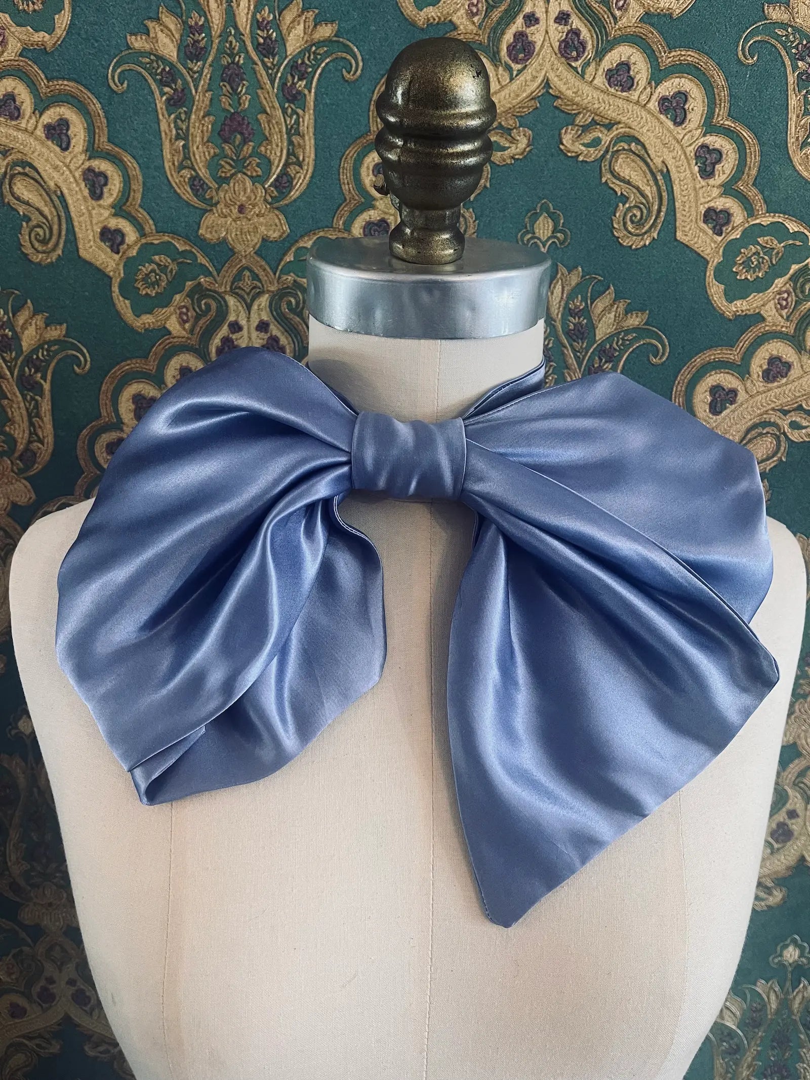 Broochella Fiocco small blue handmade  satin bow brooch. Pictured on a mannequin. The background is green and gold damask wallpaper.