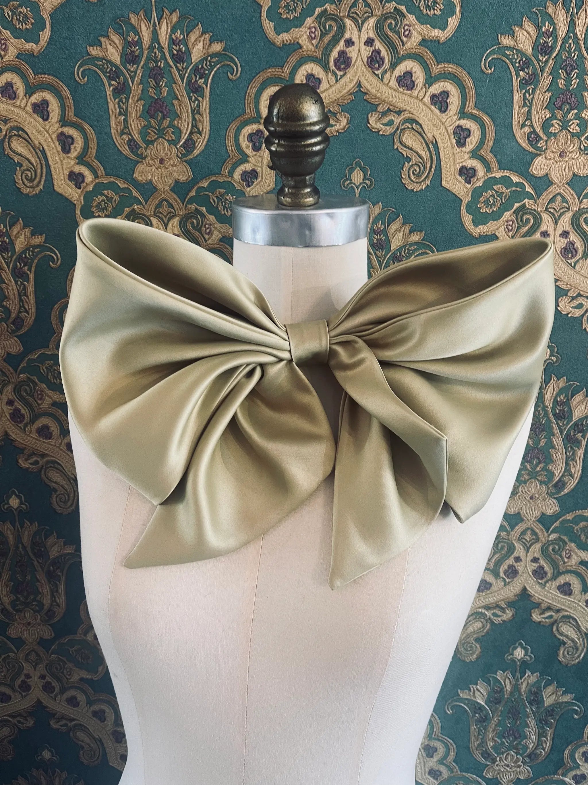 Broochella Fiocco small green handmade  satin bow brooch. Pictured on a mannequin. The background is green and gold damask wallpaper.