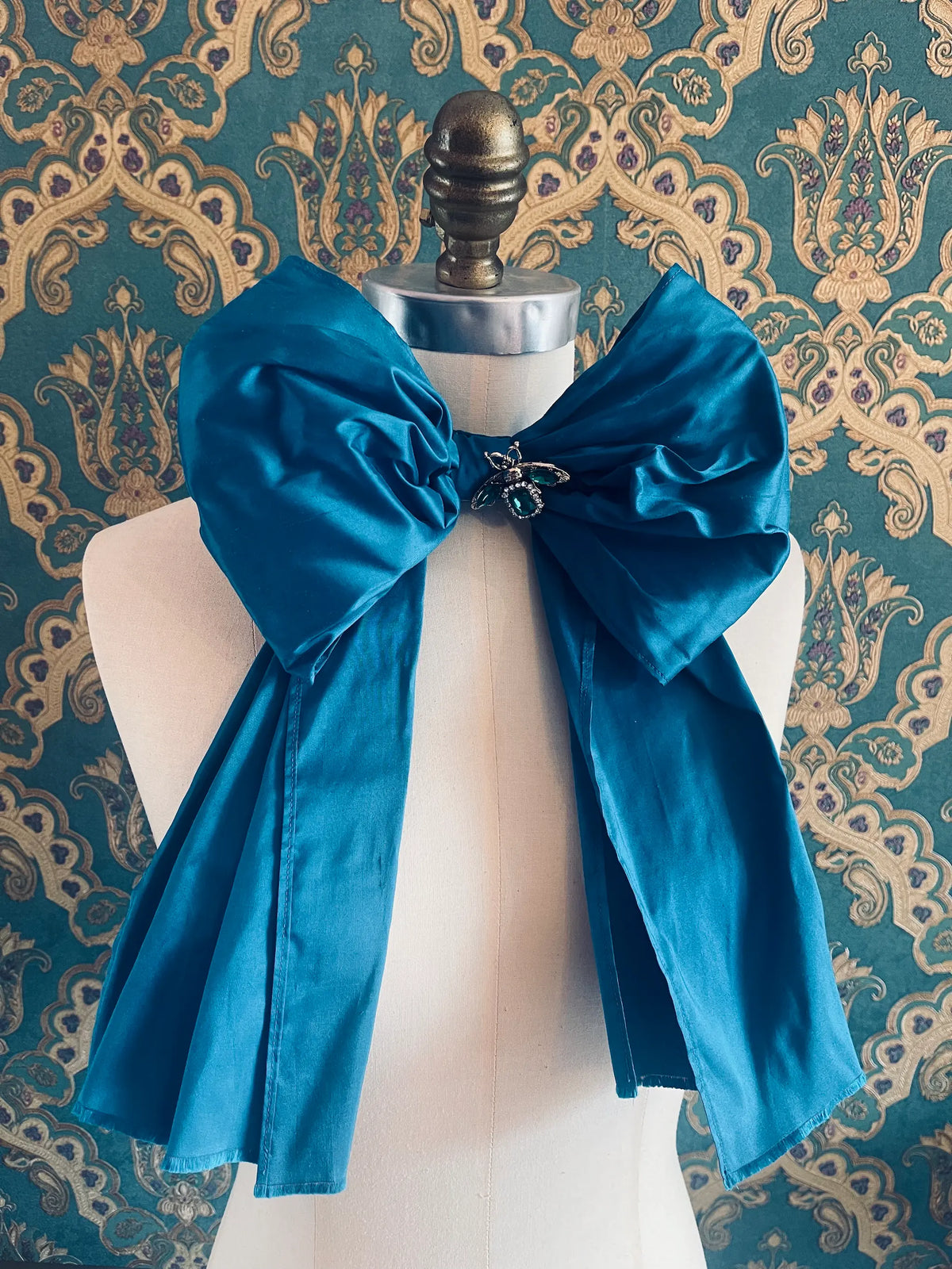 Broochella Fiocco large handmade silk embellished bow in turquoise. Pictured on a mannequin. The background is green and gold damask wallpaper.