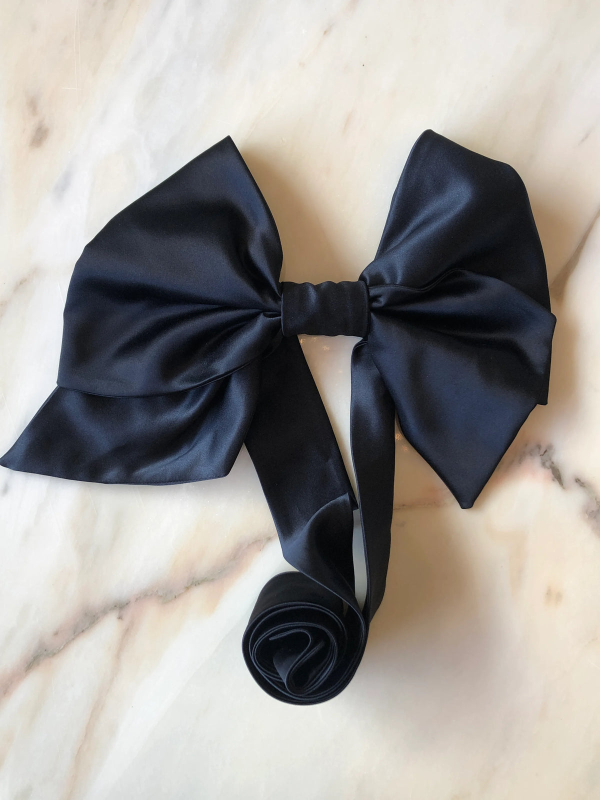 Broochella Fiocco Large Bow Brooch Pin in black satin with oversized sculptural loops and long ribbon ties, photographed on a white marble background.