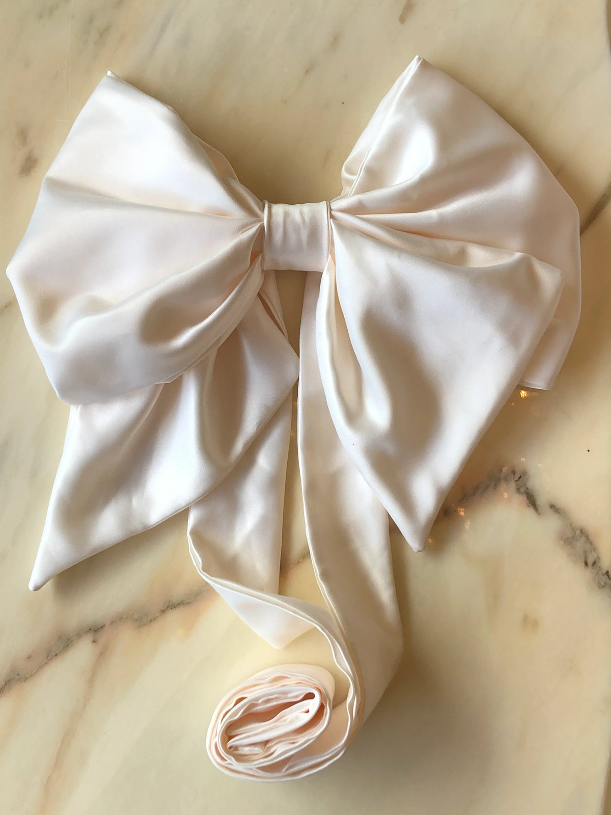 Broochella Fiocco Large Bow Brooch Pin in lustrous ivory satin with oversized sculptural loops and long ribbon ties, photographed on a white marble background.