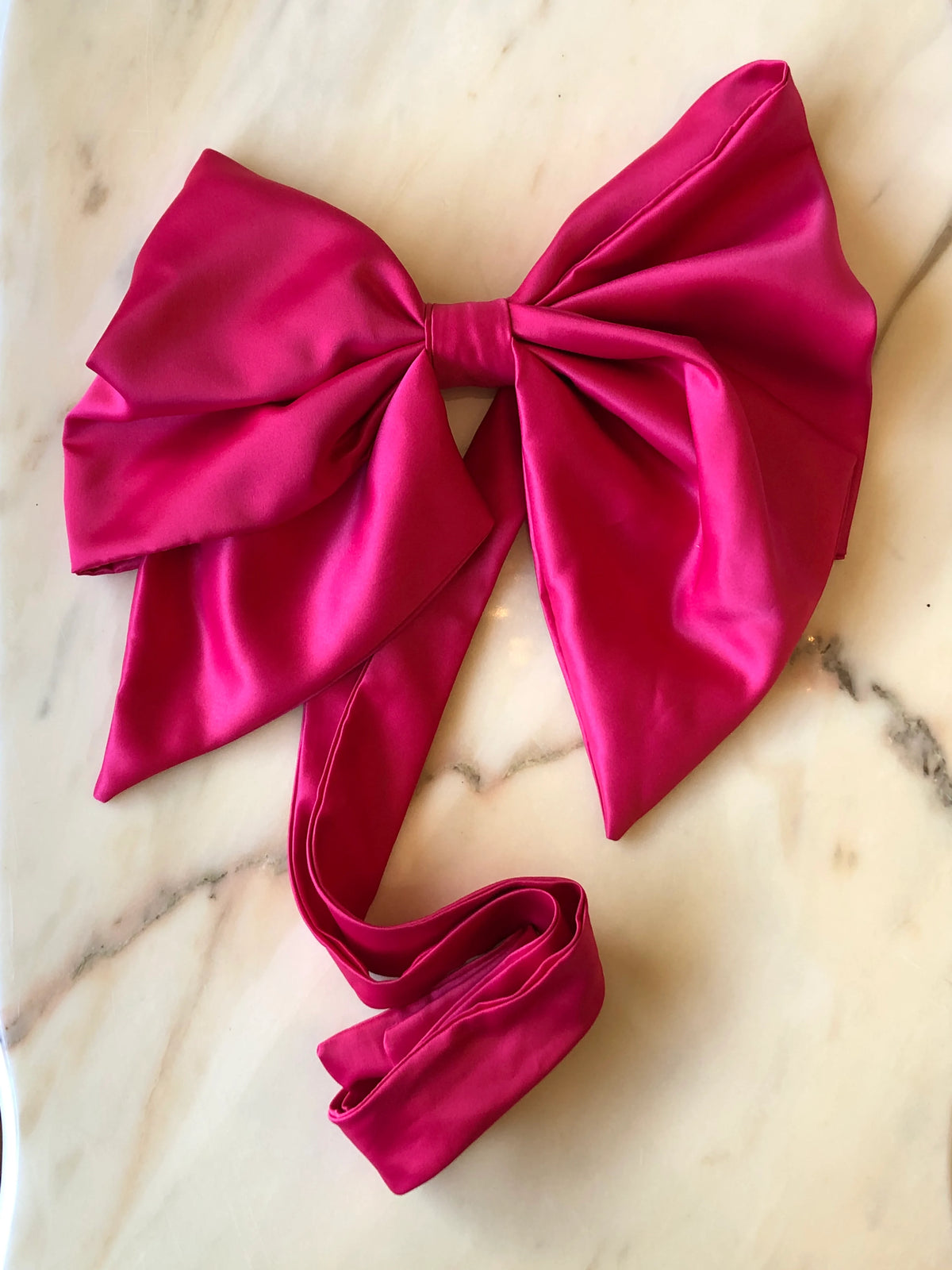 Broochella Fiocco Large Bow Brooch Pin in vibrant pink satin with oversized sculptural loops and long ribbon ties, photographed on a white marble background.