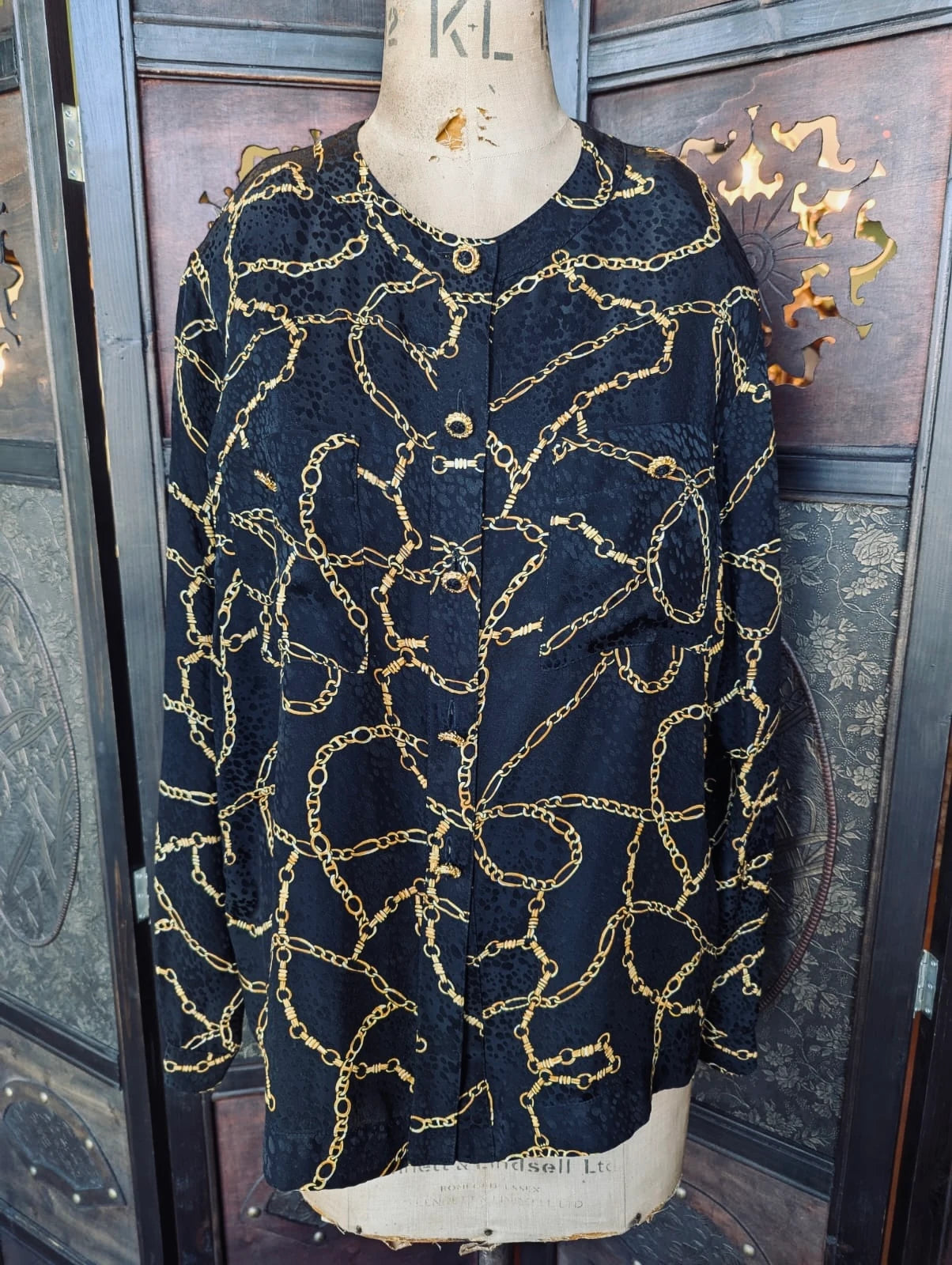 Fiona Andrews Vintage Silk Blouse – AU 10 / Vintage 14. Black long sleeve blouse with gold chain print. It is pictured on a mannequin. The background is an ornate, dark wood room divider. 