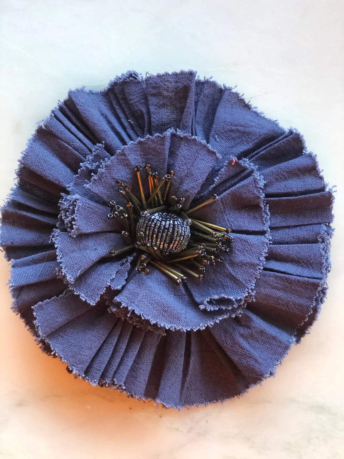 Broochella Fiordaliso Flower Brooch Pin in dusky blue, featuring pleated fabric petals with softly frayed edges and an intricate metallic black beaded stamen centre on a cream marble background.