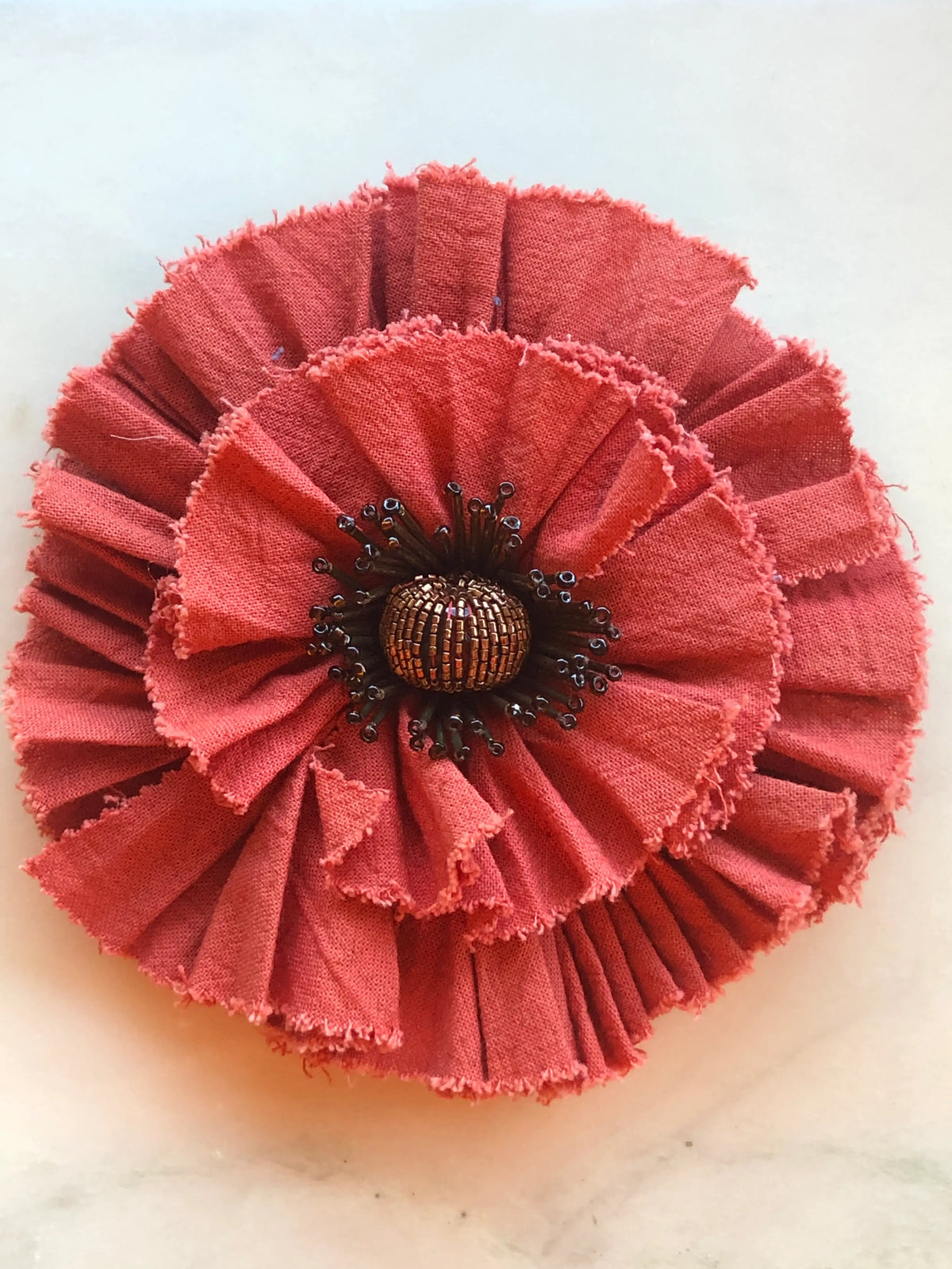 Broochella Fiordaliso Flower Brooch Pin in warm red, featuring pleated fabric petals with softly frayed edges and an intricate bronze and black beaded stamen centre on a cream marble background.