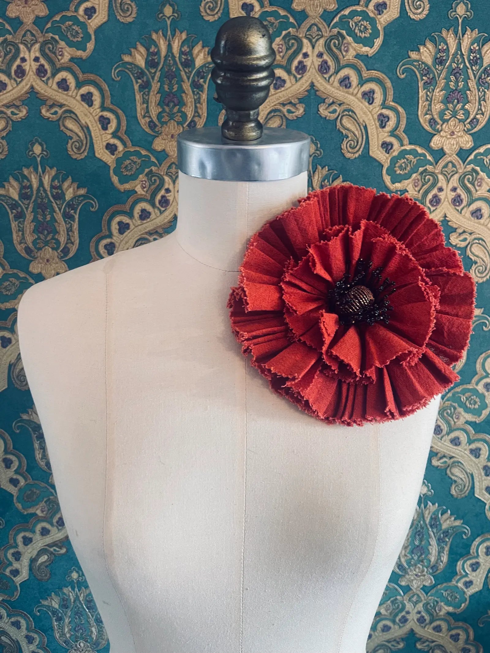 Broochella fiordaliso handmade large fabric flower brooch in red. Styled on a mannequin. The background is green and gold damask wallpaper