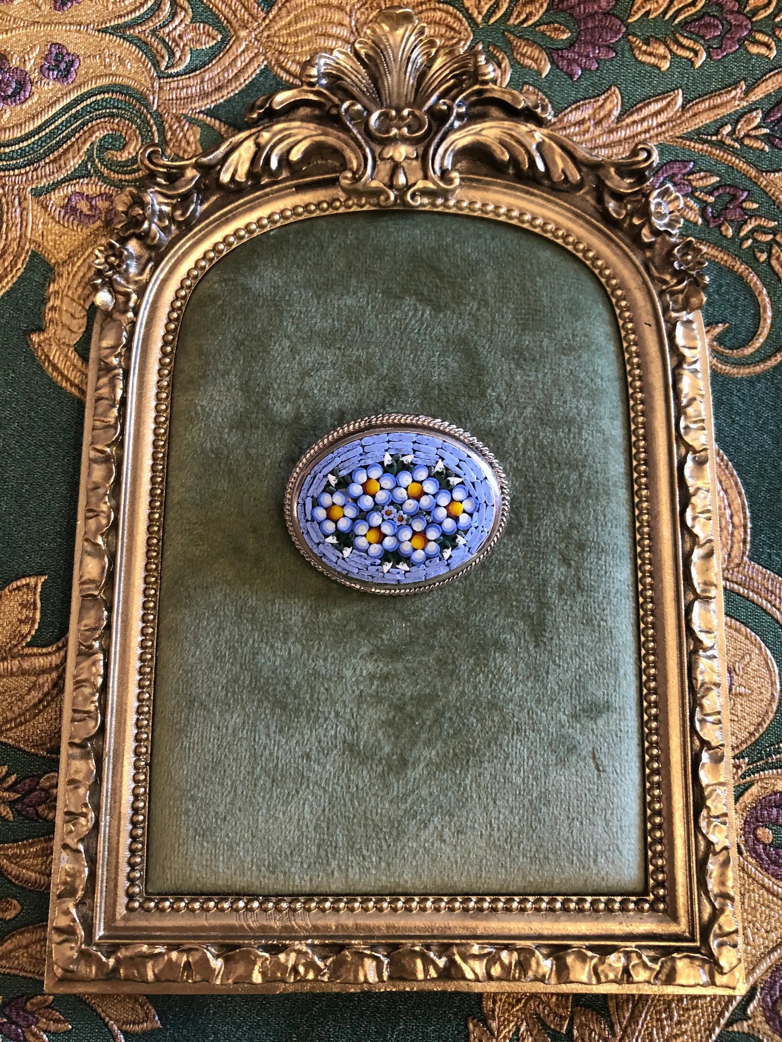 A Broochella Fiorella  3cm x 4cm vintage Italian micro mosaic oval brooch featuring a hand assembled glass tessera design of forget-me-not flowers in cornflower blue, white and yellow on a blue ground, set within a silver-tone frame with twisted rope edge detail, photographed on a green velvet tray, with a gold frame. The background is green and gold damask wallpaper.